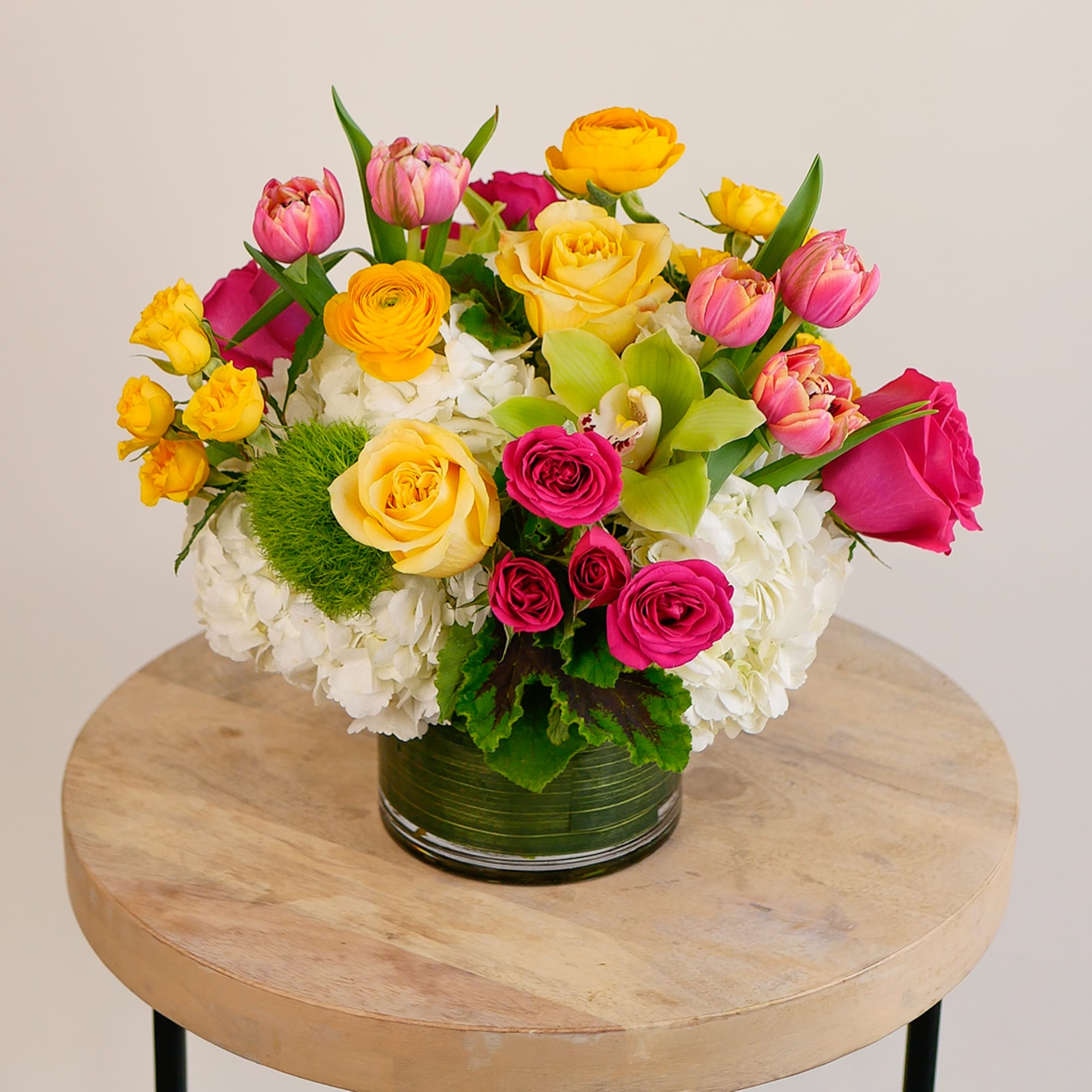 Low arrangement of yellow and pink roses, tulips, orchids and white hydrangeas in a clear glass vase