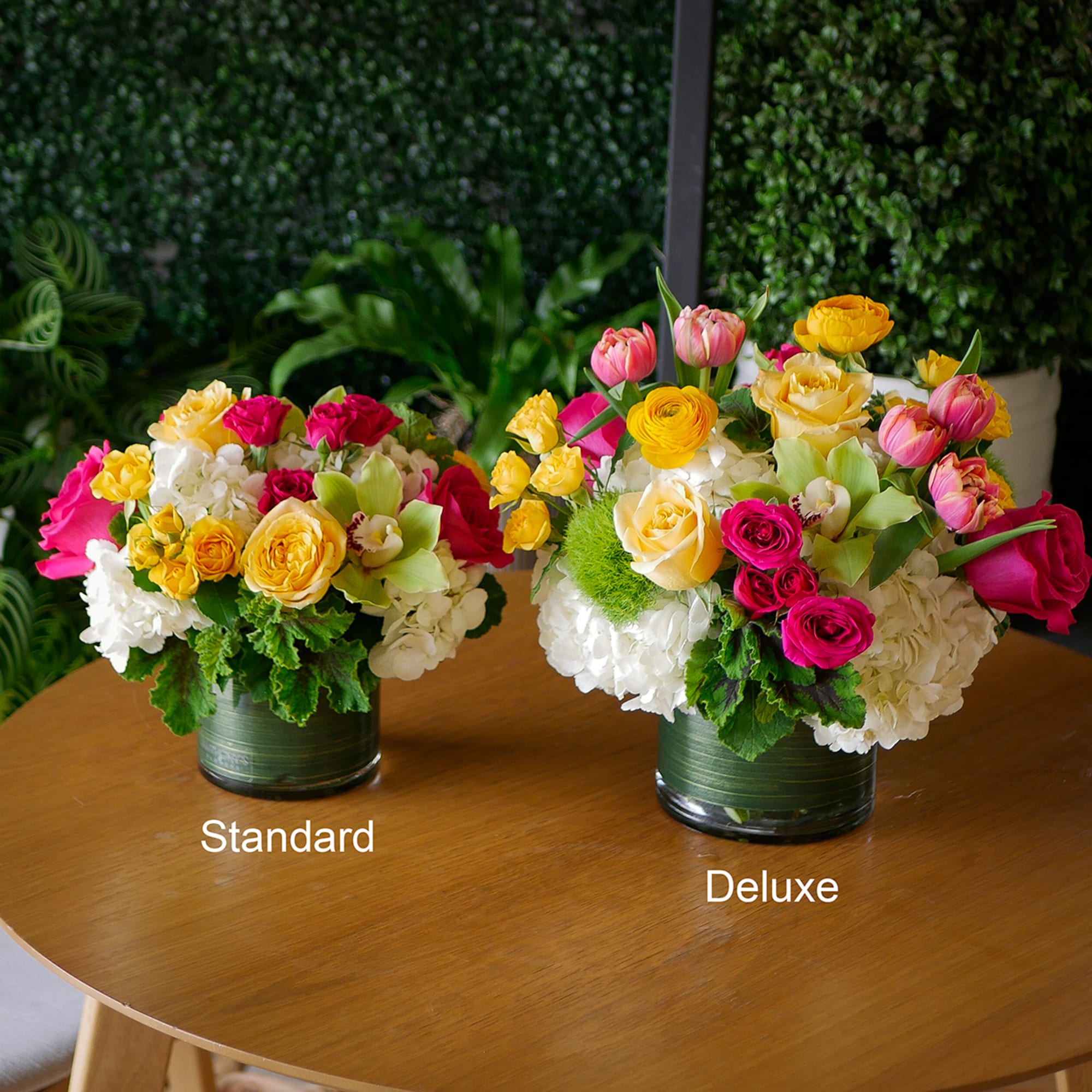 Two bright mixed arrangements of roses and tulips in leaf-lined glass vases labeled standard and deluxe on a table