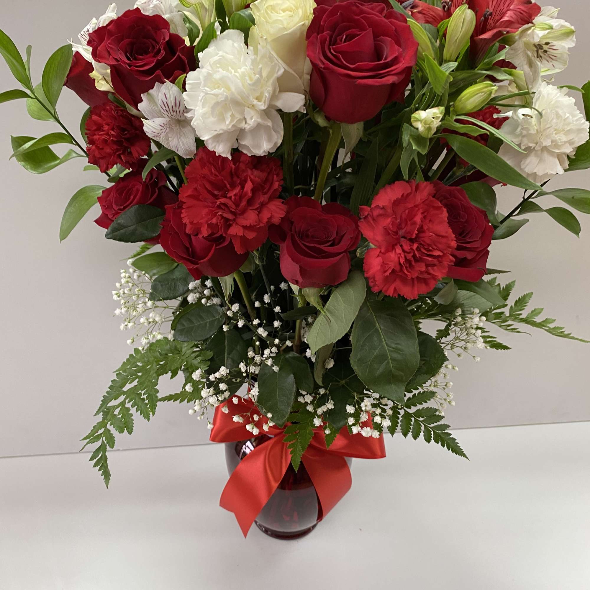 Arrangement of red roses, white blooms, and red carnations in a red vase with a bow