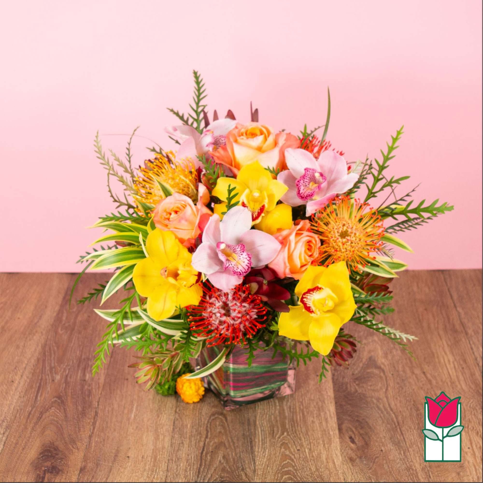 Bright arrangement of yellow orchids, pink roses, and orange pincushion protea in a clear square glass vase