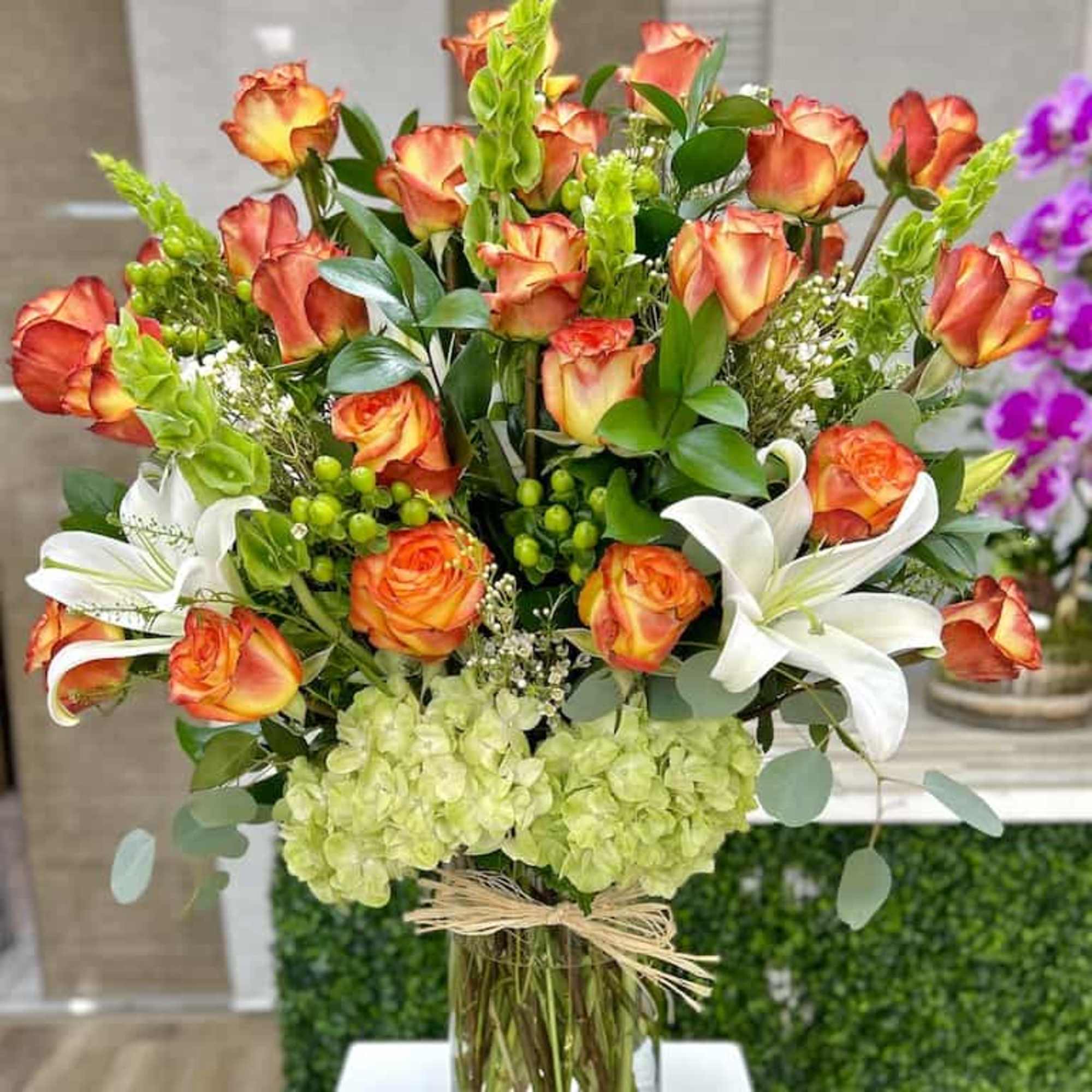 Tall arrangement of orange roses, white lilies, and green hydrangeas in a clear glass vase