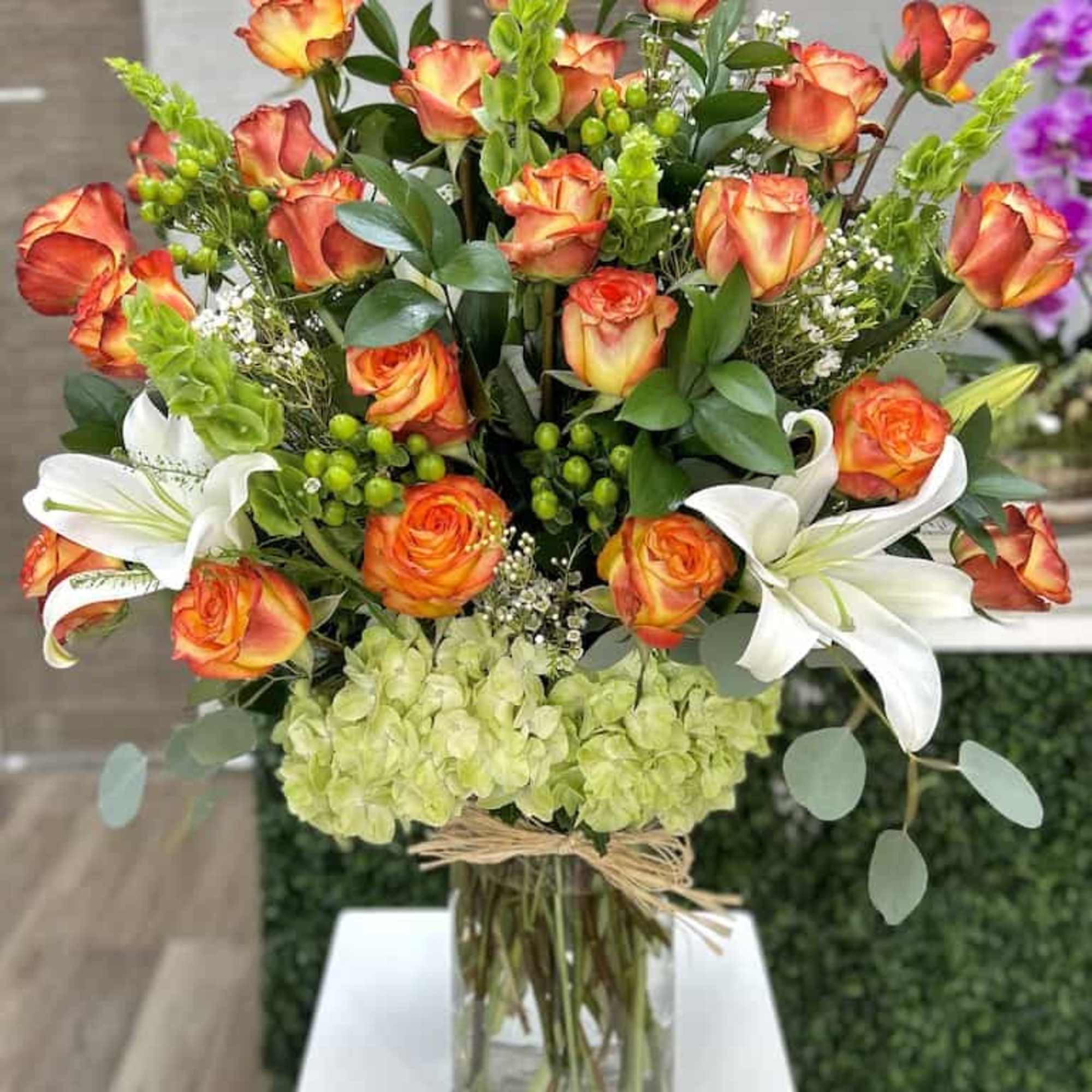 Tall arrangement of orange roses, white lilies, and green hydrangeas in a clear glass vase