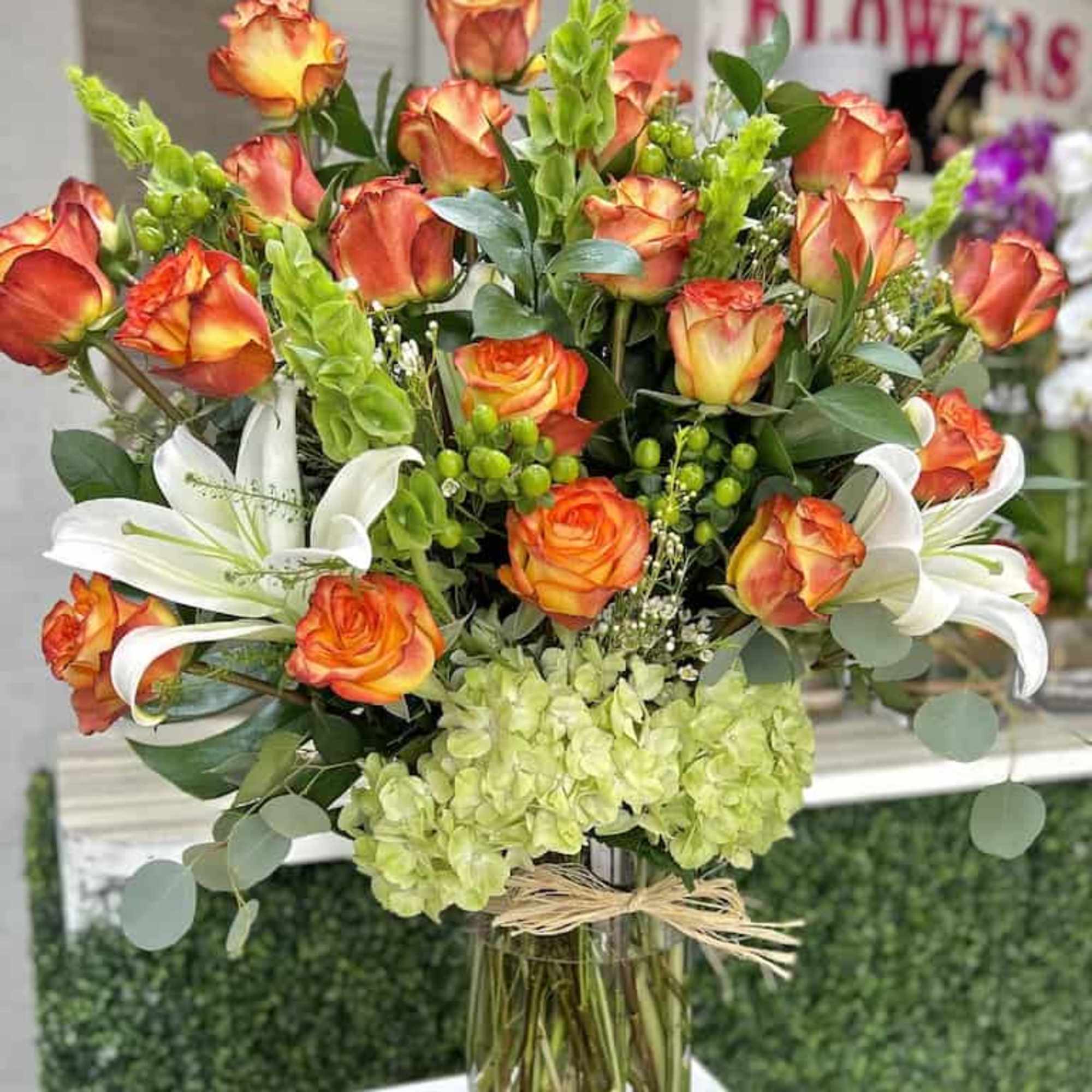 Tall arrangement of orange roses, white lilies, and green hydrangeas in a clear glass vase