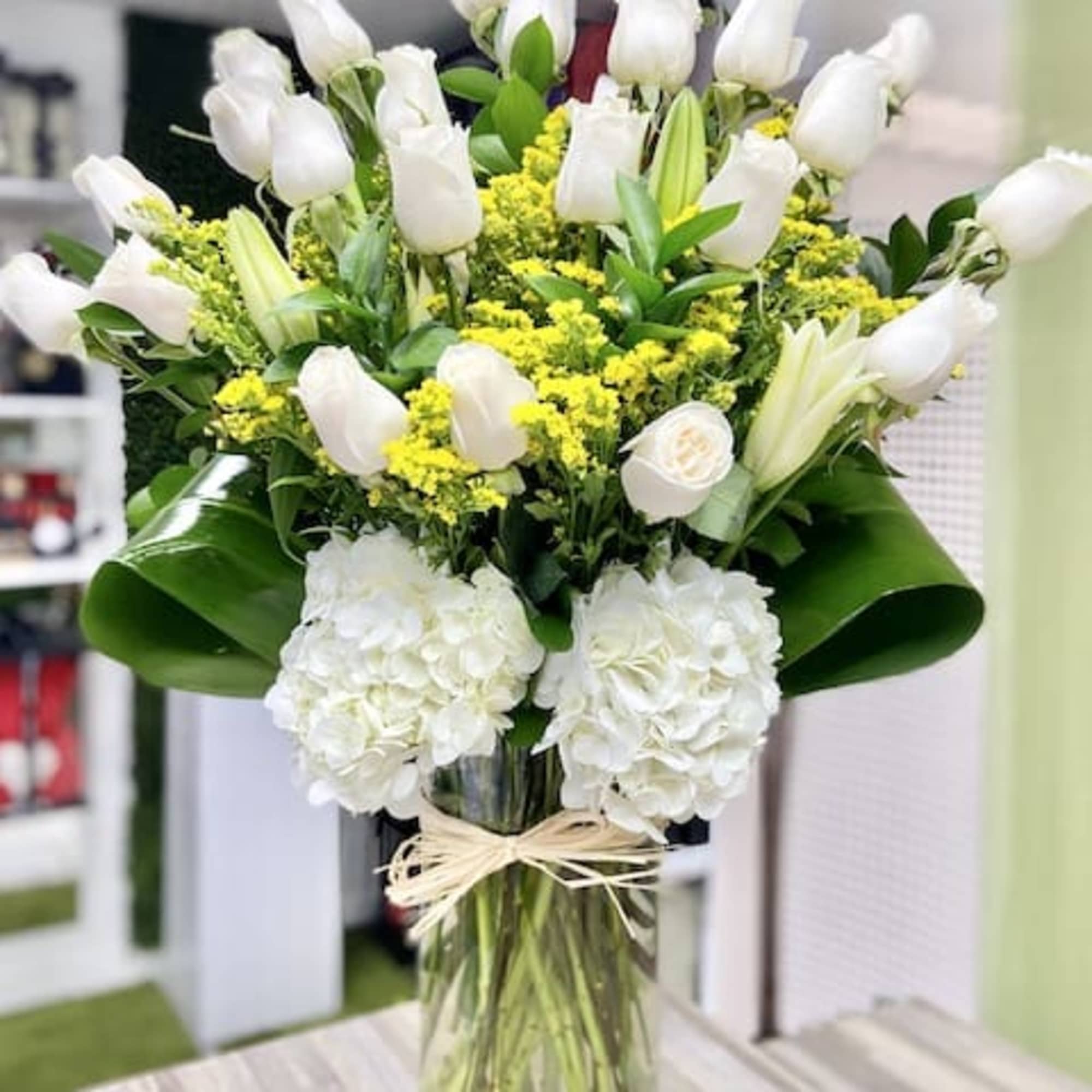 Tall arrangement of white roses, lilies, and hydrangeas with yellow accents in a clear glass vase