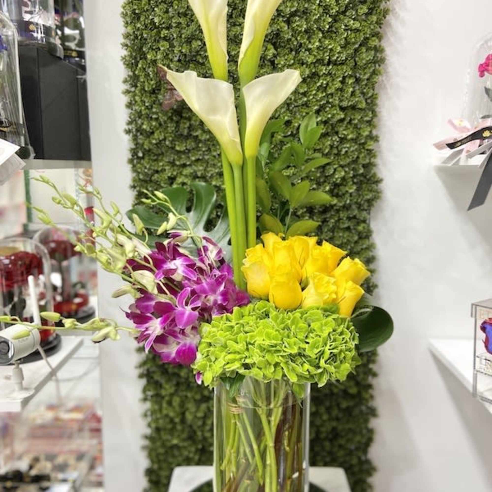 Tall glass vase arrangement with white calla lilies, yellow roses, purple orchids, and green hydrangea