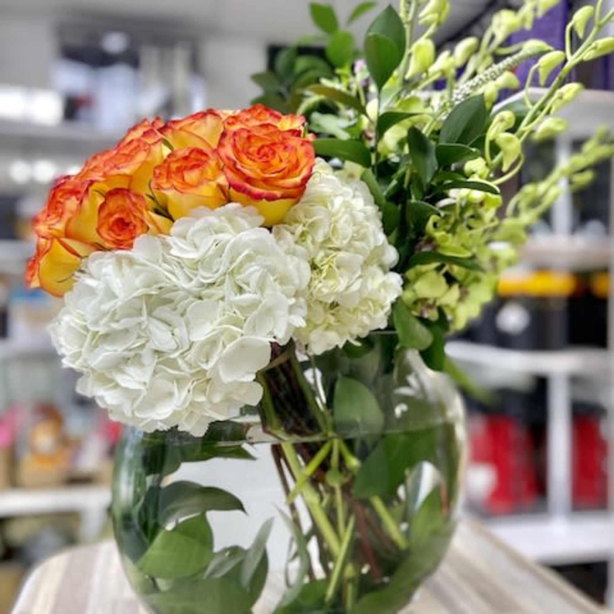 Round glass vase with white hydrangeas, orange-yellow roses, and green orchid stems arranged asymmetrically