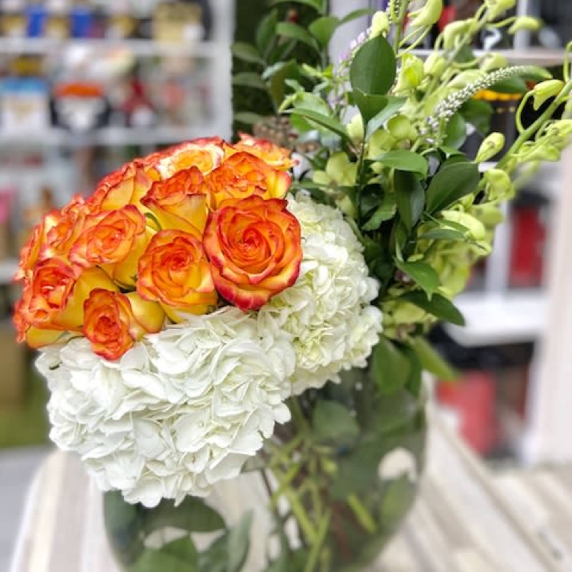 Round glass vase of orange-yellow roses, white hydrangeas, and tall green buds.