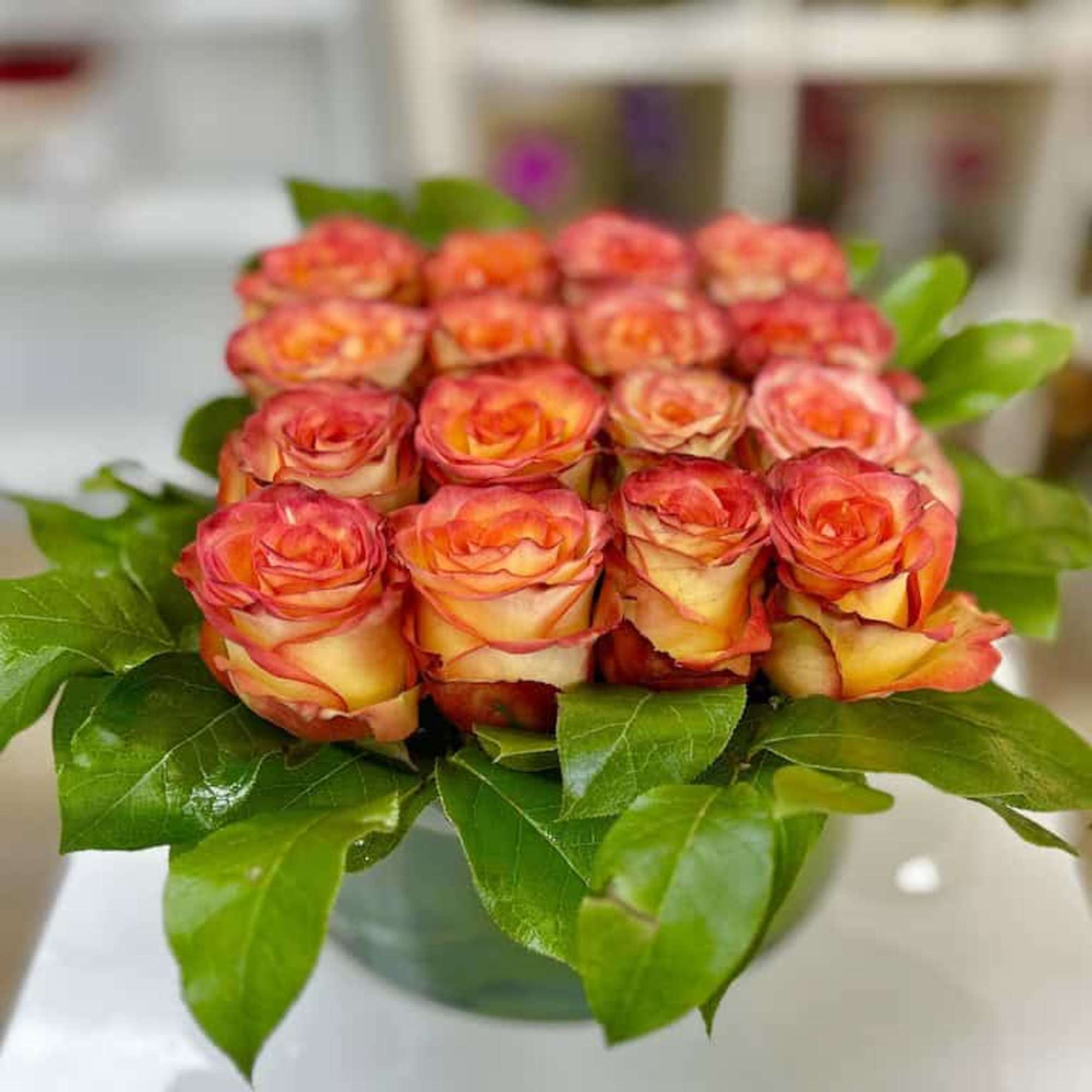 Compact arrangement of orange and yellow roses framed by glossy green leaves in a low glass vase