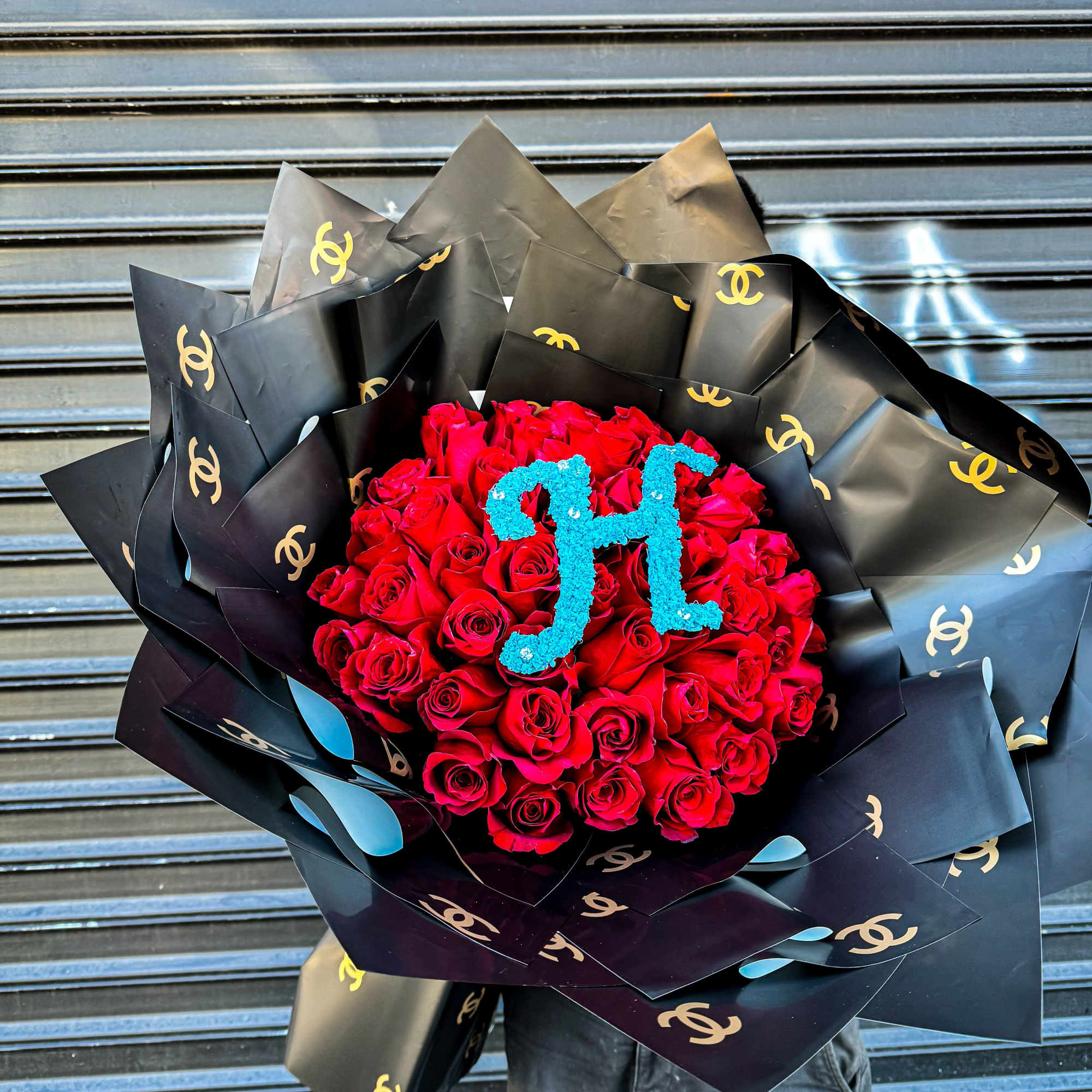 Large bouquet of red roses in black logo wrap with teal floral initials on top.
