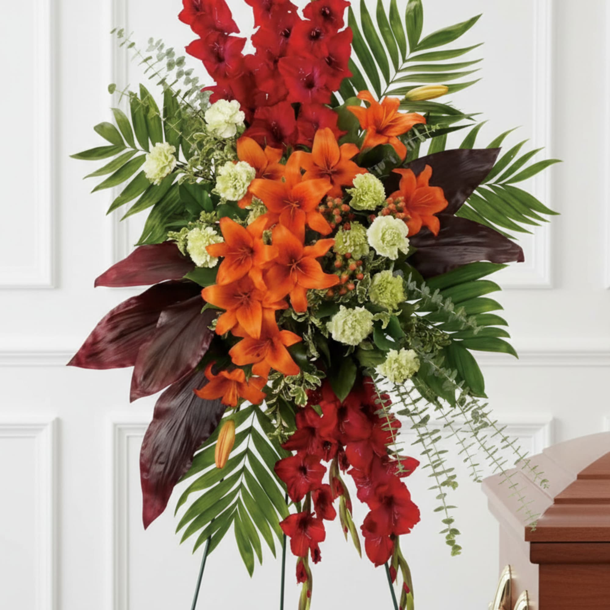 Tall standing spray of red gladiolus, orange lilies, and green carnations on an easel beside a casket.