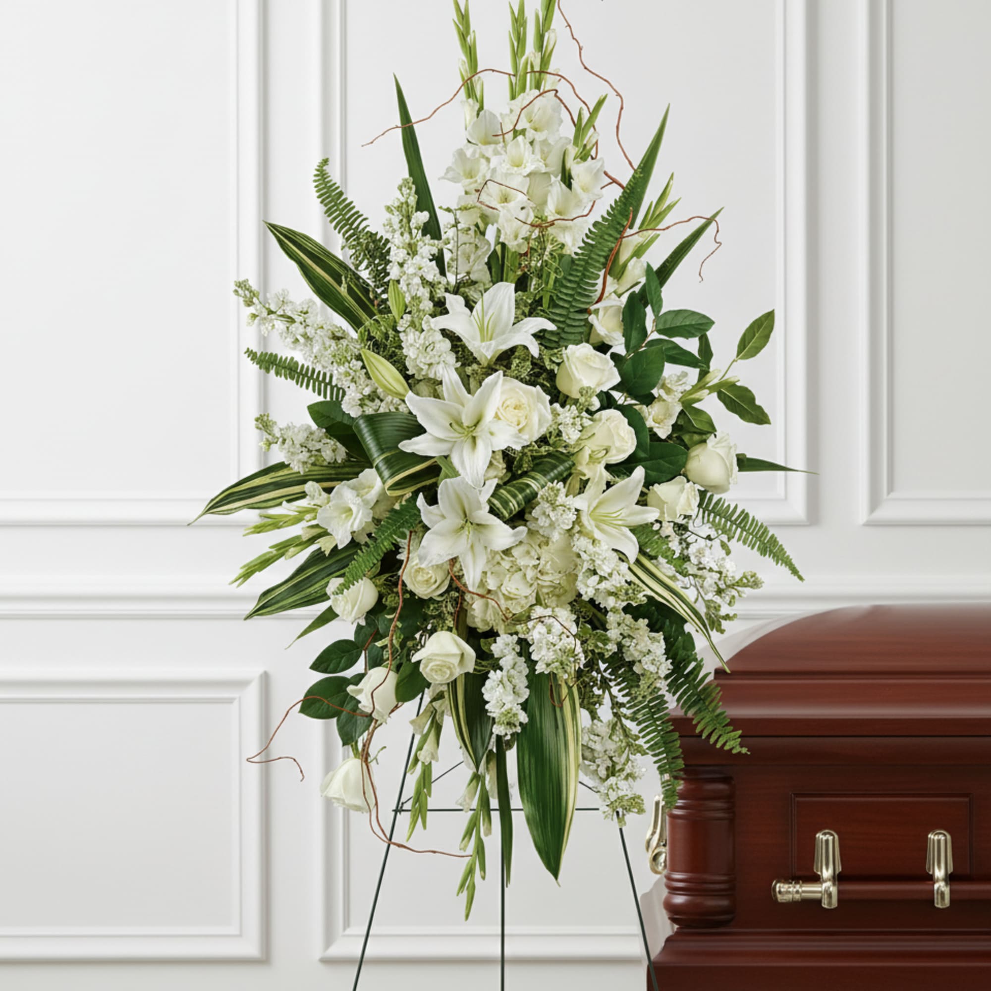 Tall white lily and rose standing spray on an easel beside a wood casket
