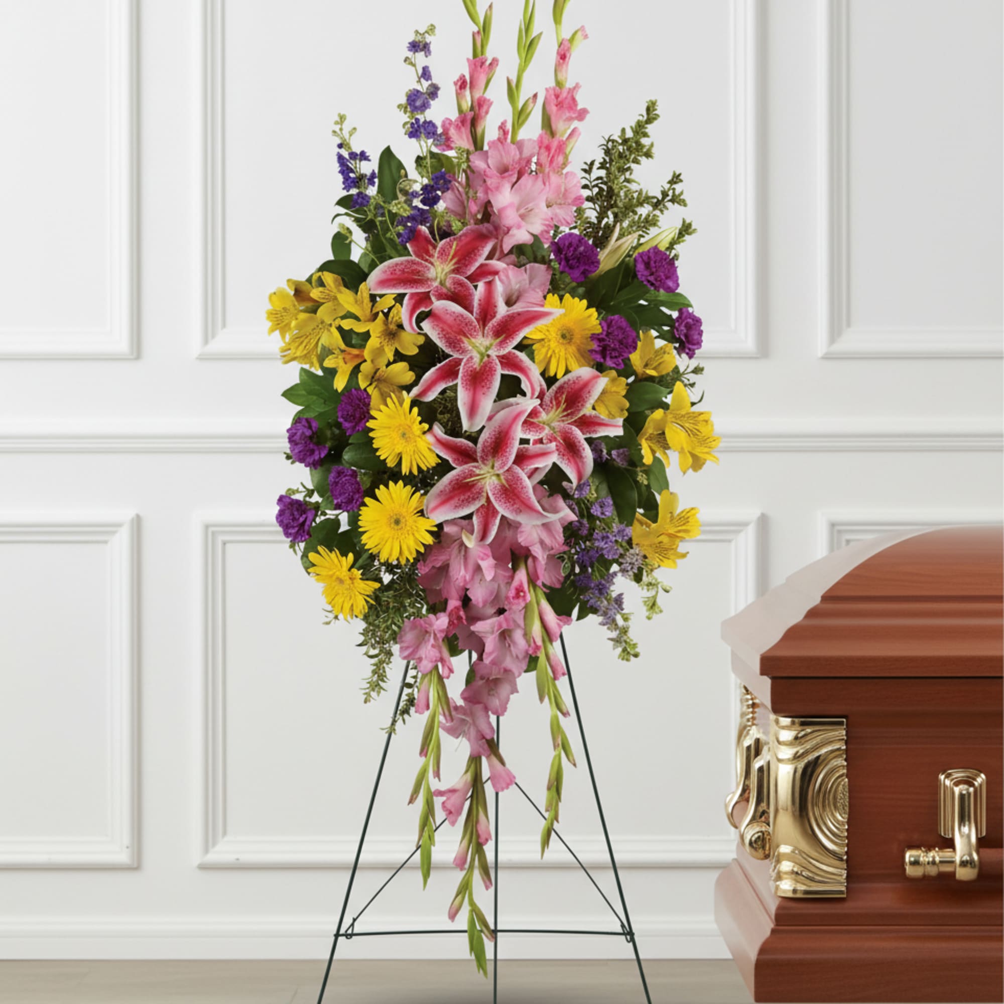Tall standing spray of pink lilies, yellow daisies, and purple flowers on an easel beside a wooden casket