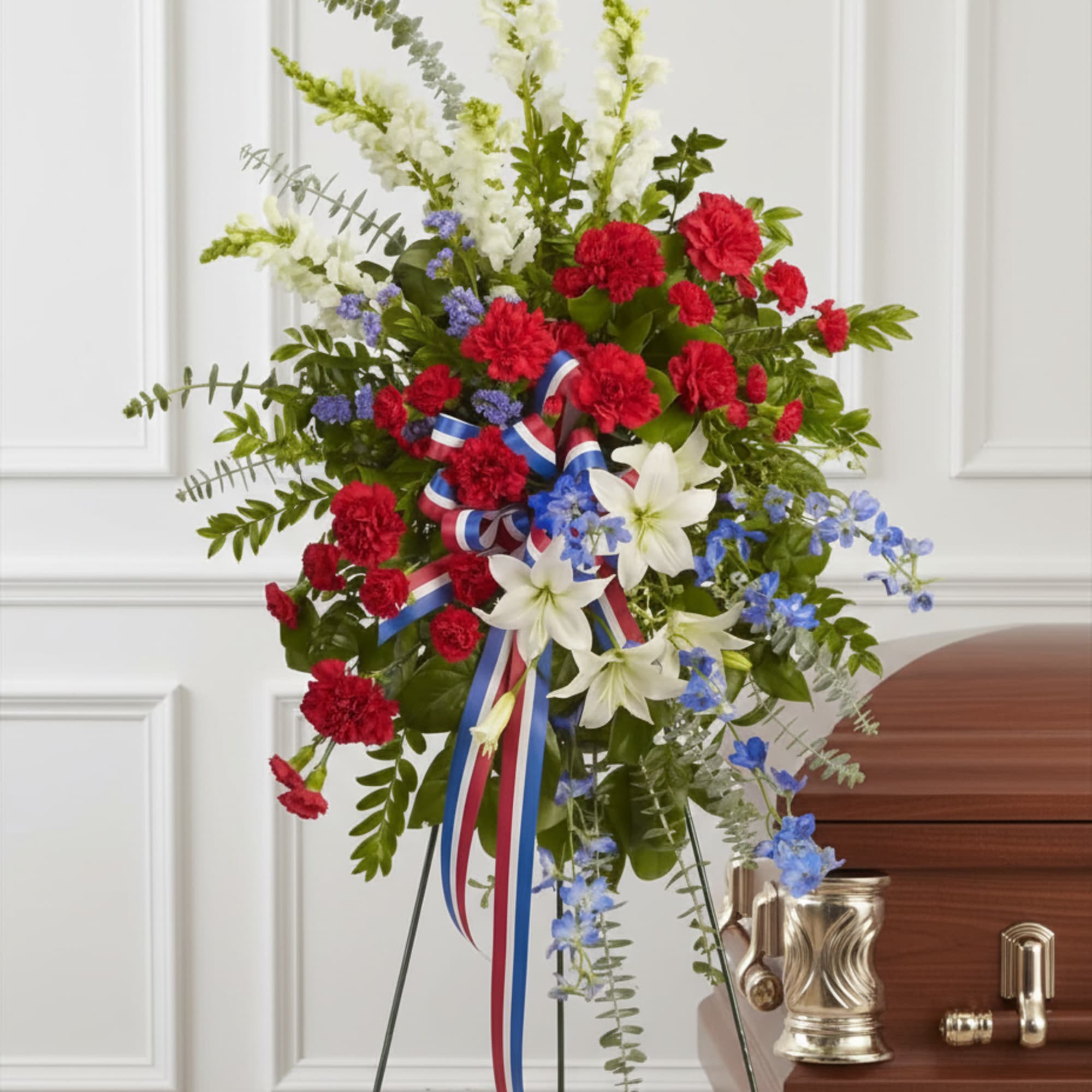 Tall red, white, and blue standing spray with carnations, lilies, and ribbon on an easel beside a casket.
