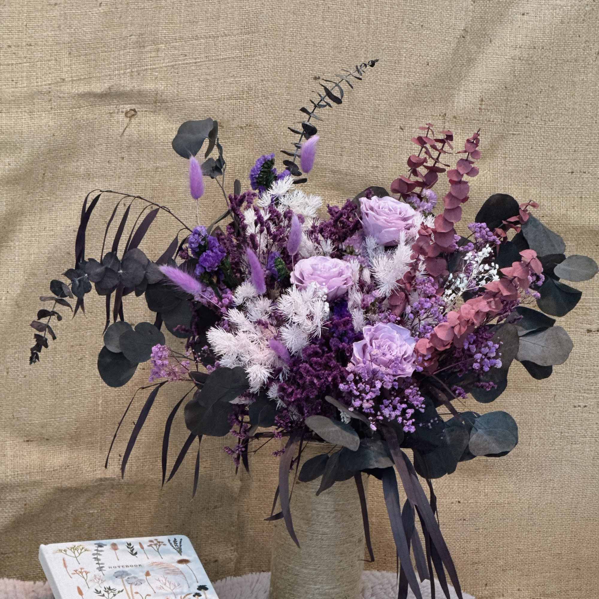 Tall arrangement of purple and lavender flowers in a twine-wrapped vase beside a floral notebook.