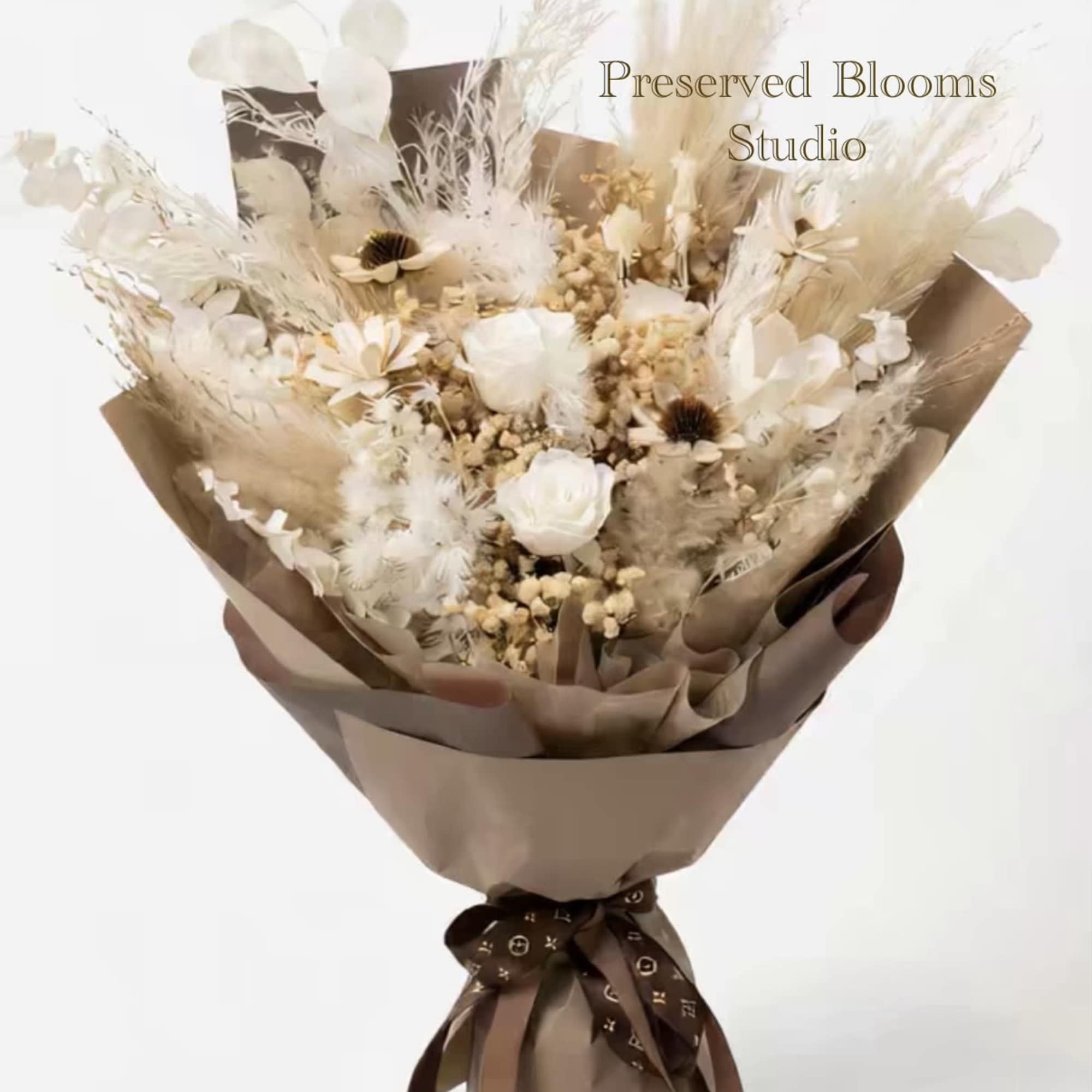 Neutral-toned bouquet of preserved white roses and dried flowers wrapped in brown paper with a dark ribbon.