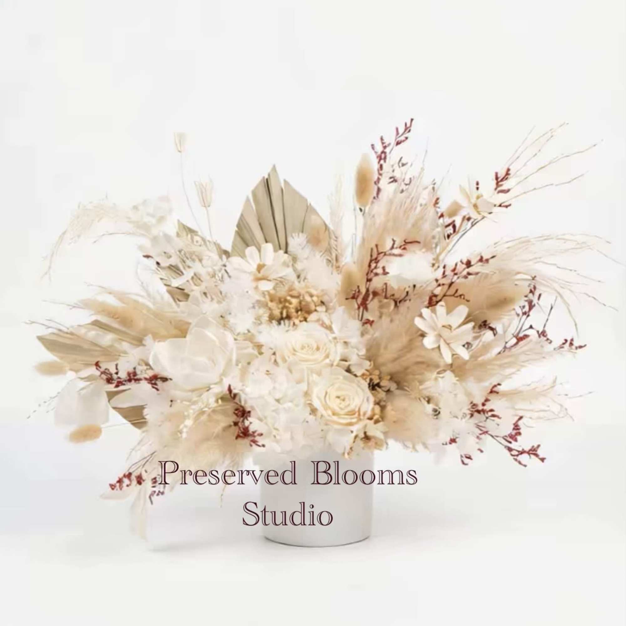 Low arrangement of preserved neutral-toned flowers with roses and dried grasses in a white cylinder vase.
