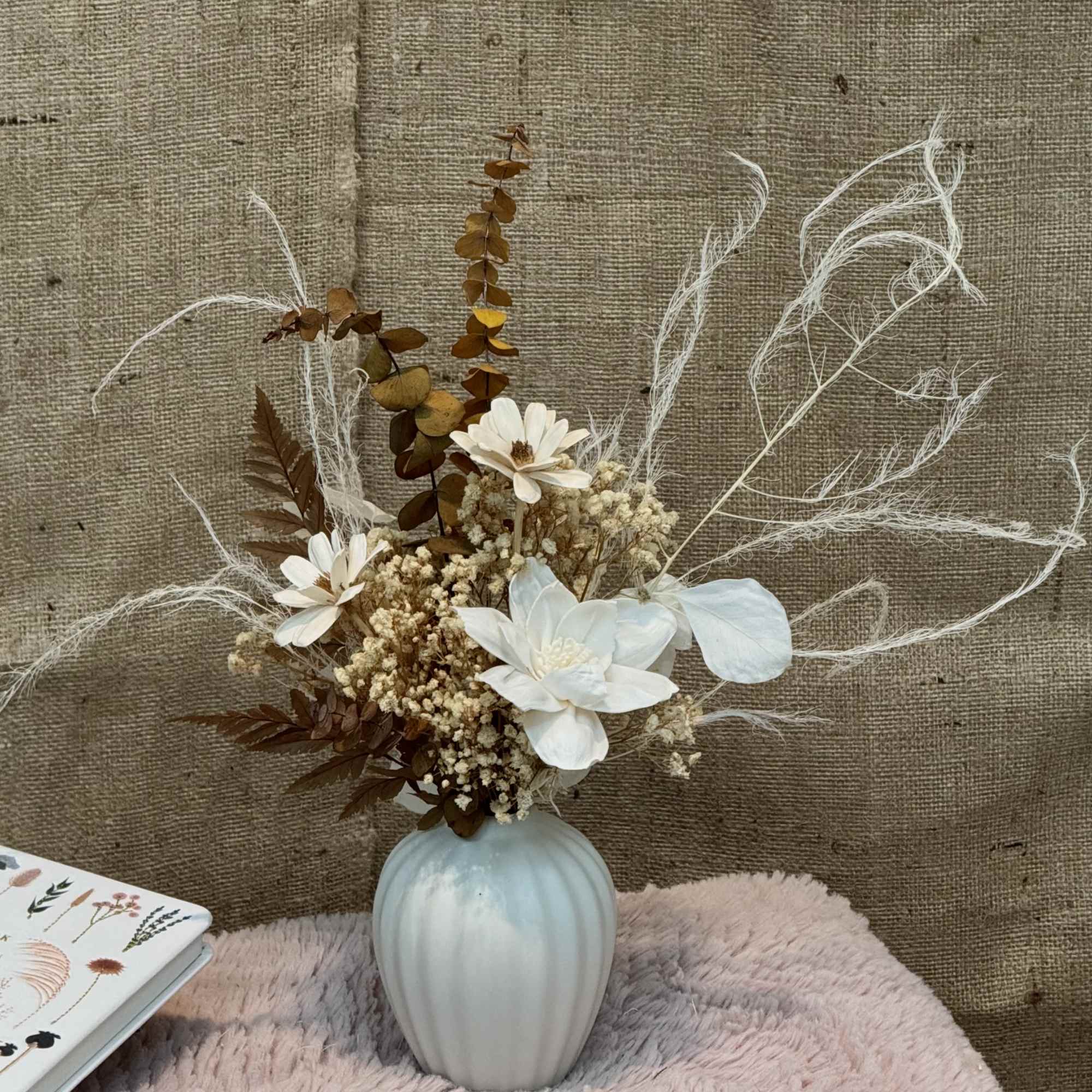 Small arrangement of white and beige dried flowers in a ribbed white vase on a soft pink surface.