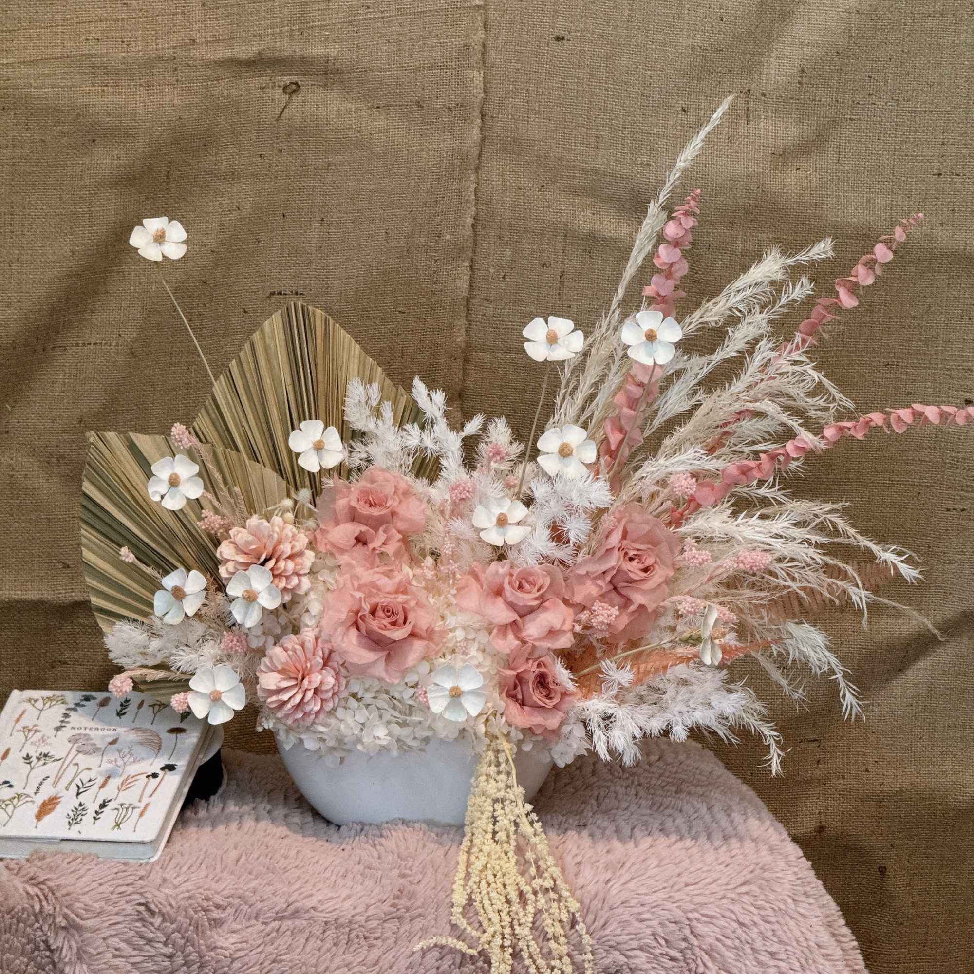 Asymmetrical arrangement of pink roses and white blooms with dried palm and feathery accents in a white bowl