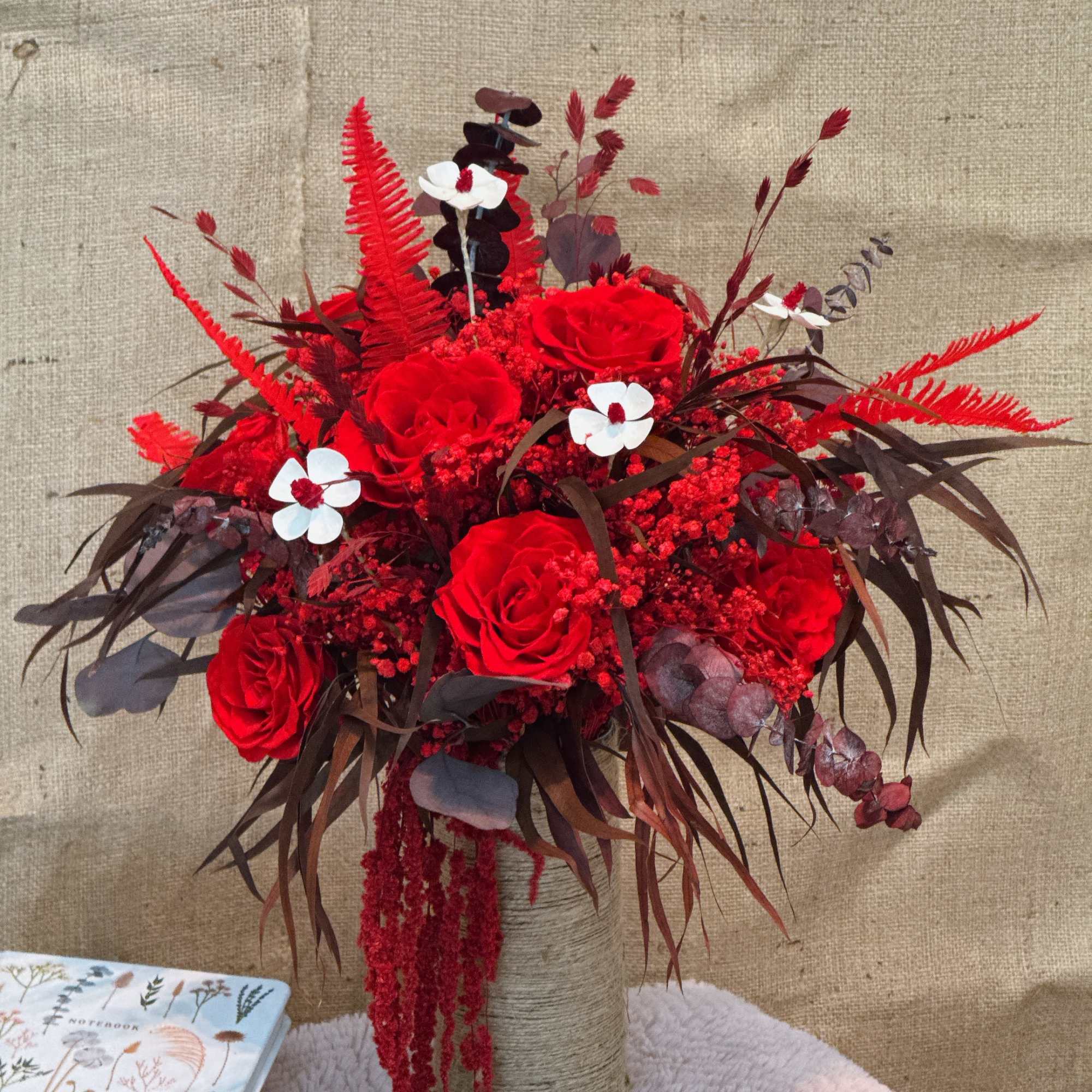 Tall arrangement of red roses with red accents and small white blooms in a twine-wrapped vase.
