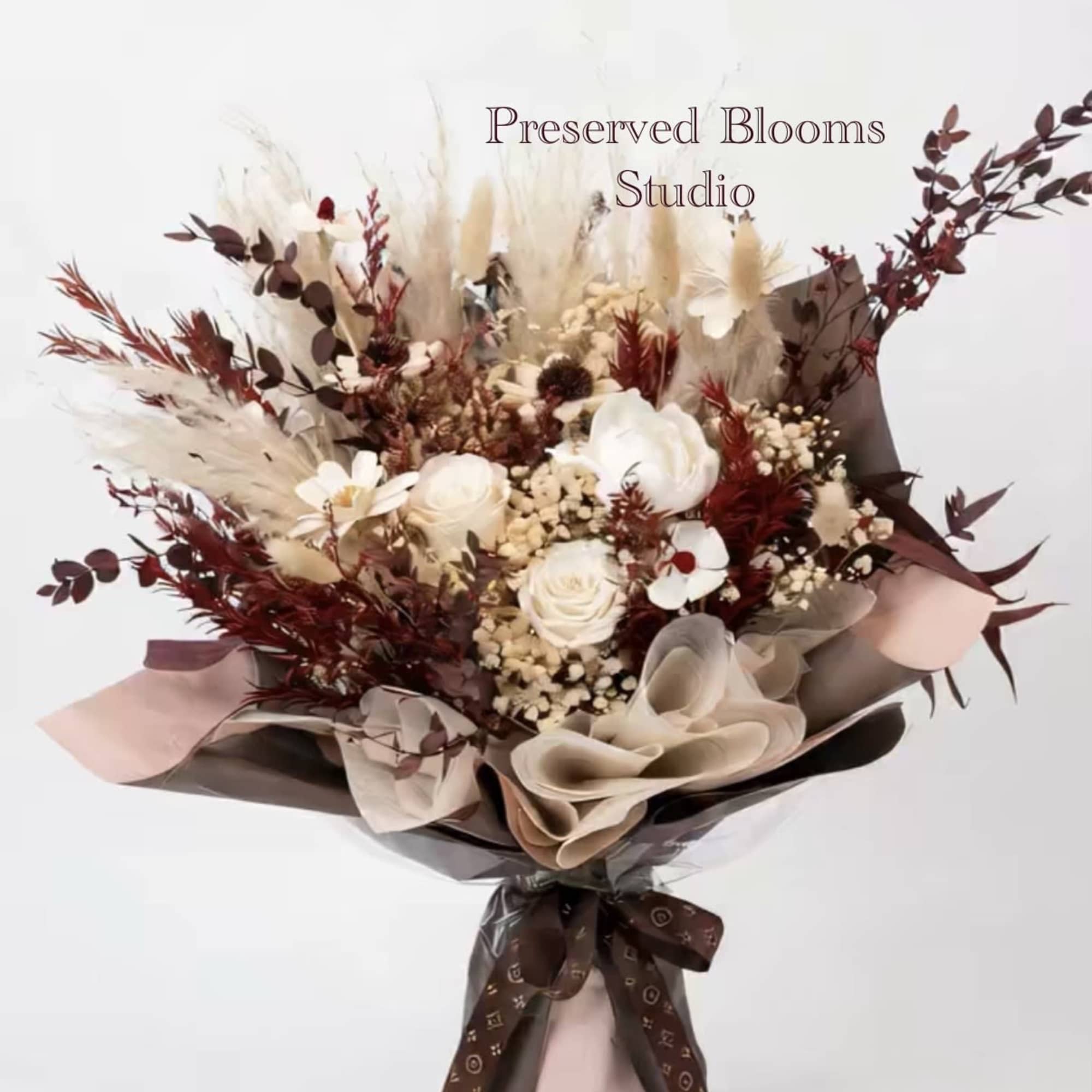 Bouquet of preserved white roses with burgundy dried foliage wrapped in brown and blush paper with a printed ribbon