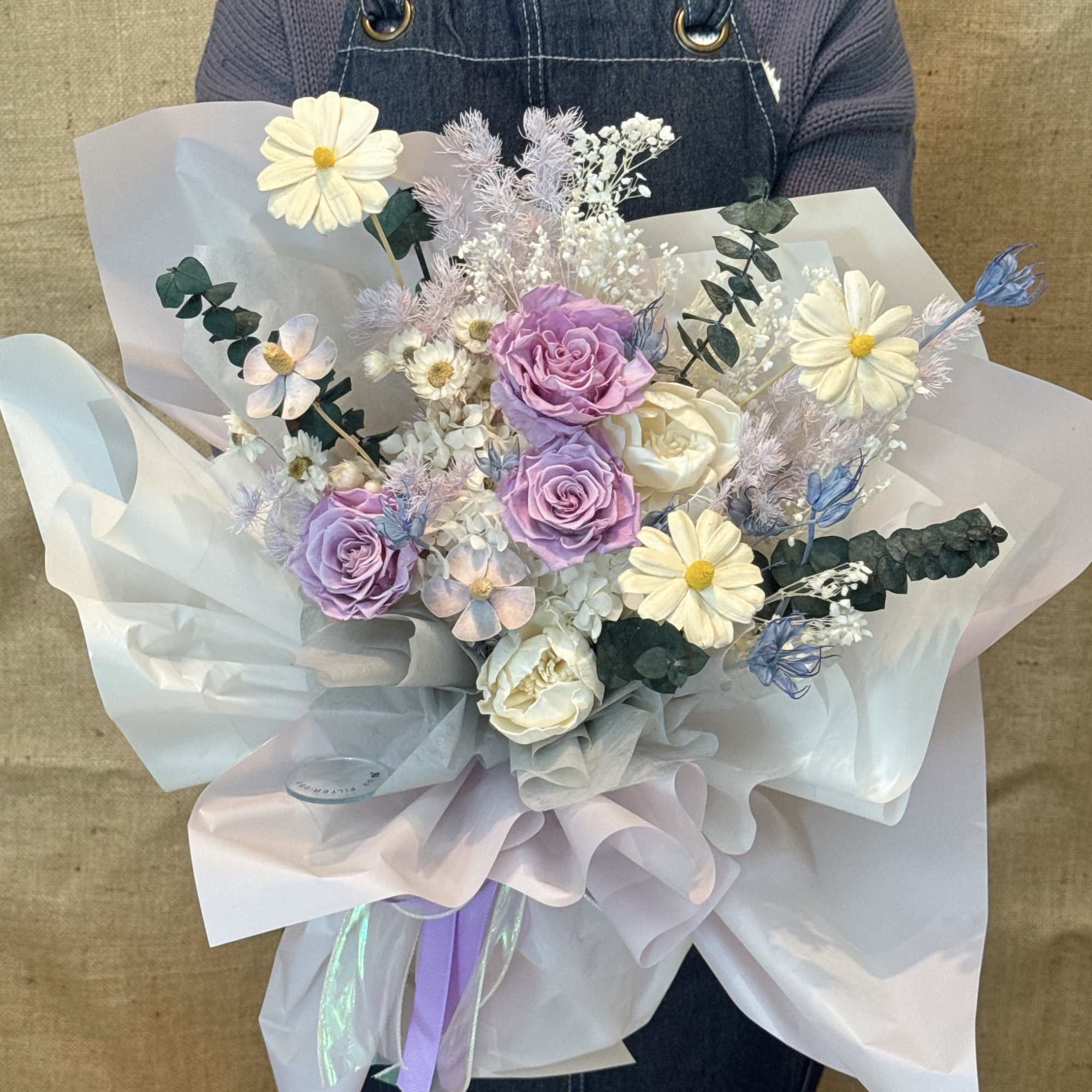 Hand-tied bouquet of lavender roses and white daisies wrapped in pale ruffled paper.