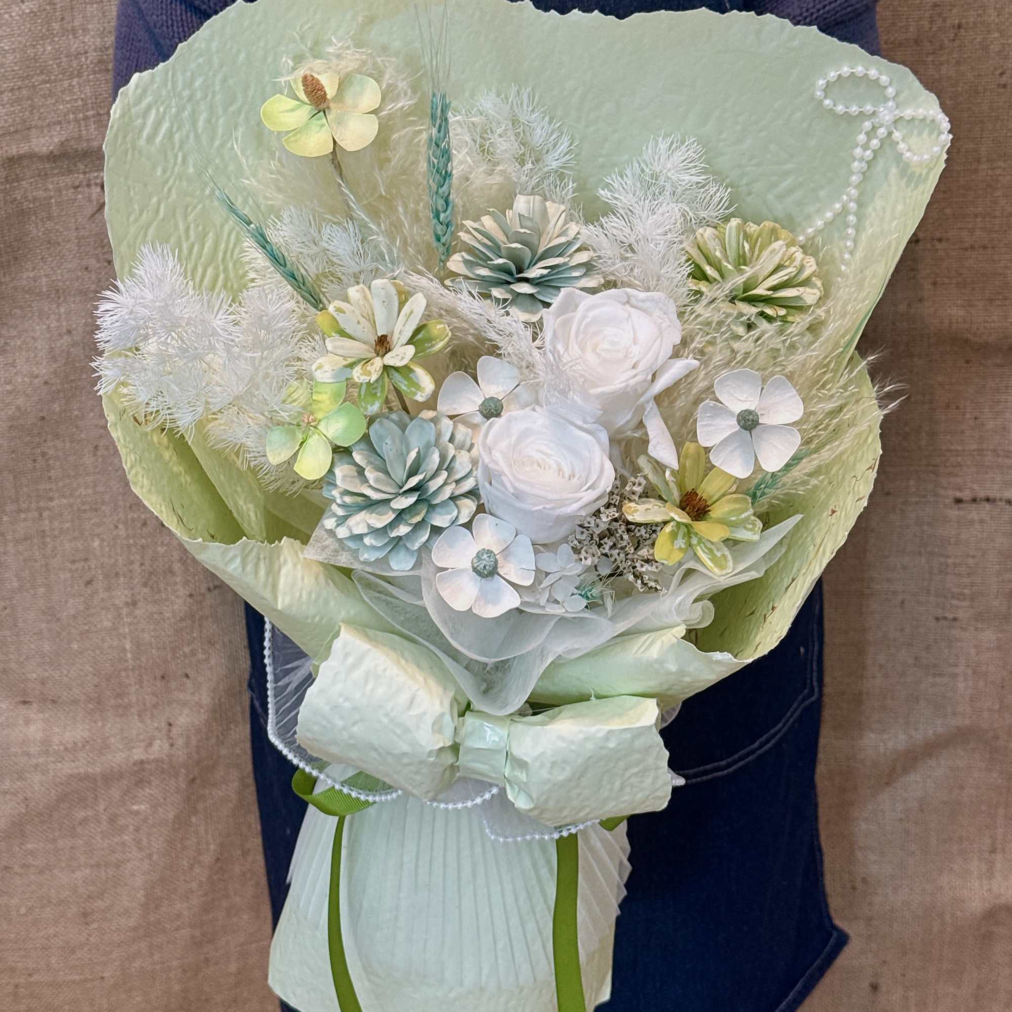 Pastel green-wrapped bouquet of white and mint artificial flowers held by a person in an apron