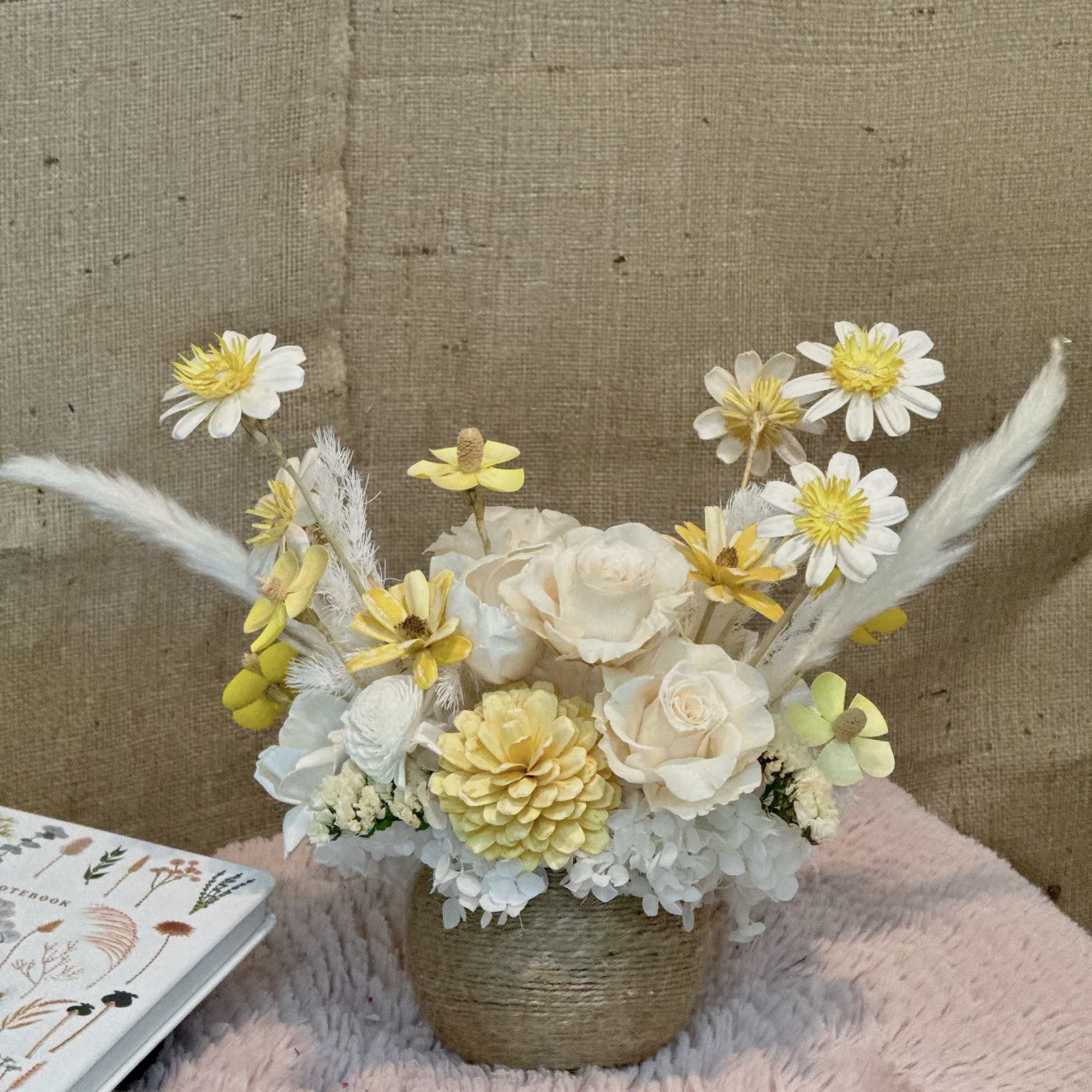 Low arrangement of cream and yellow flowers in a twine-wrapped pot on a pink textured surface