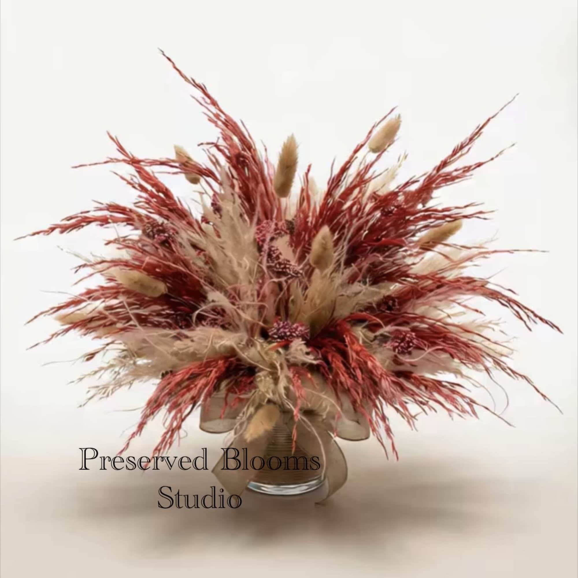 Preserved arrangement of tan and rust dried grasses and seed clusters in a clear glass vase.