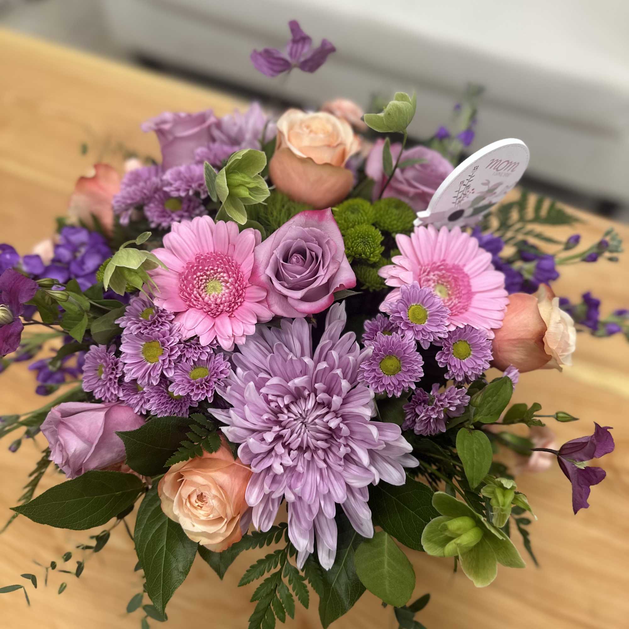 Round arrangement of pink gerberas, lavender mums, and peach roses with a small Mom card pick.