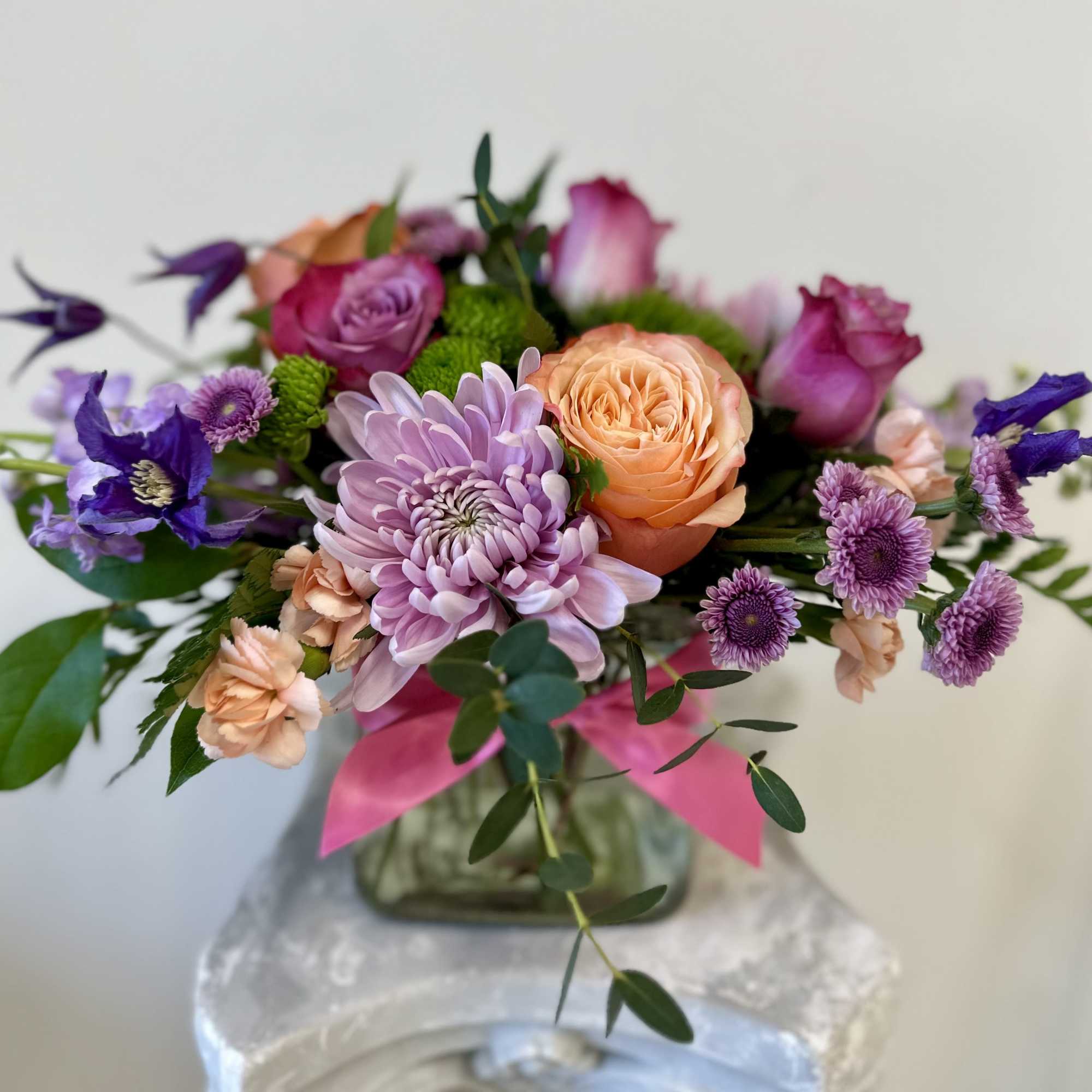 Low arrangement of purple mums, peach roses, and mixed blooms in a glass vase with a pink ribbon.