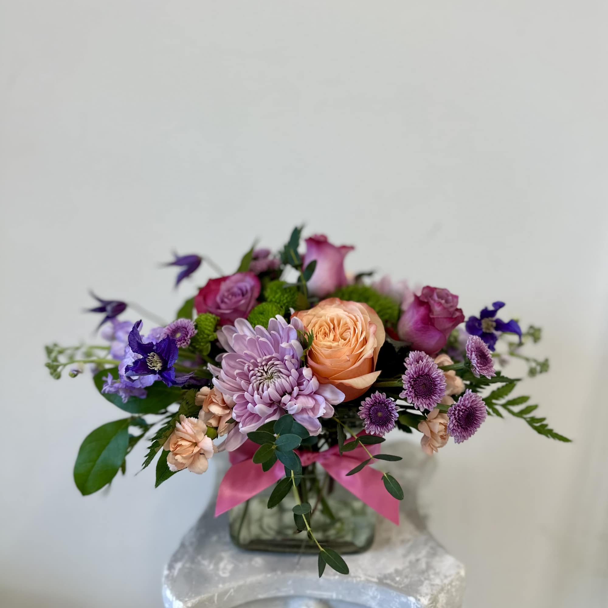 Low bouquet of peach roses, lavender mums, and purple flowers in a square glass vase with a pink ribbon.