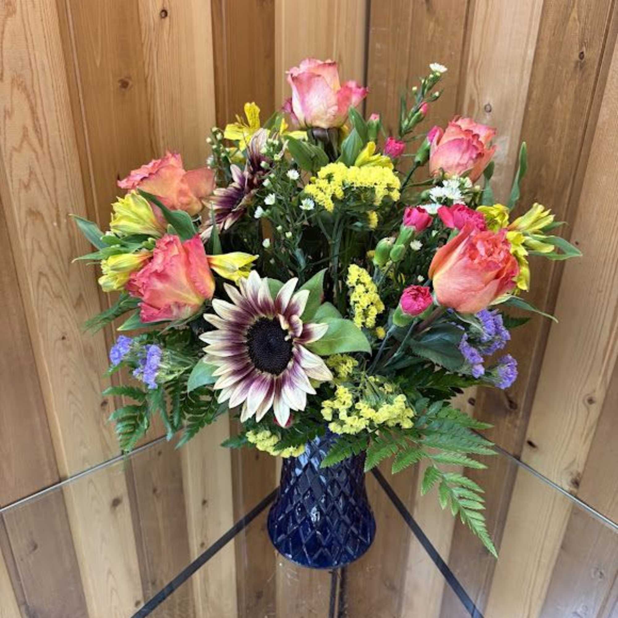 Mixed bouquet with pink roses, sunflower, and yellow blooms in a dark blue glass vase