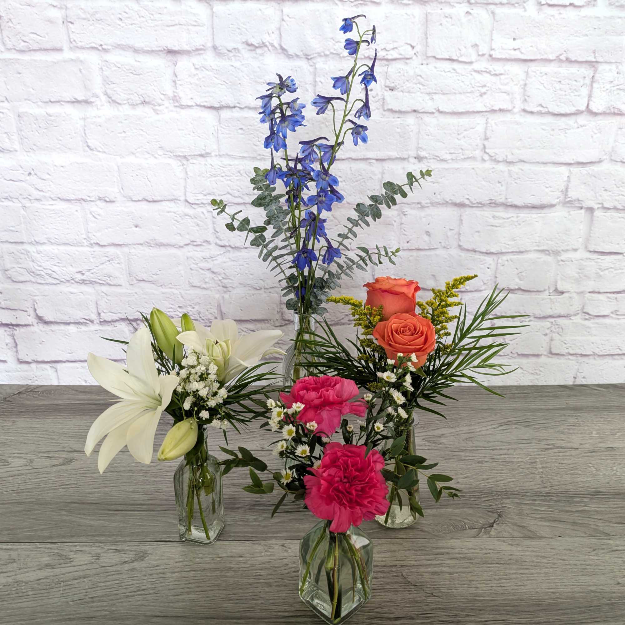 Three small floral arrangements in glass vases with lilies, roses, and blue delphinium