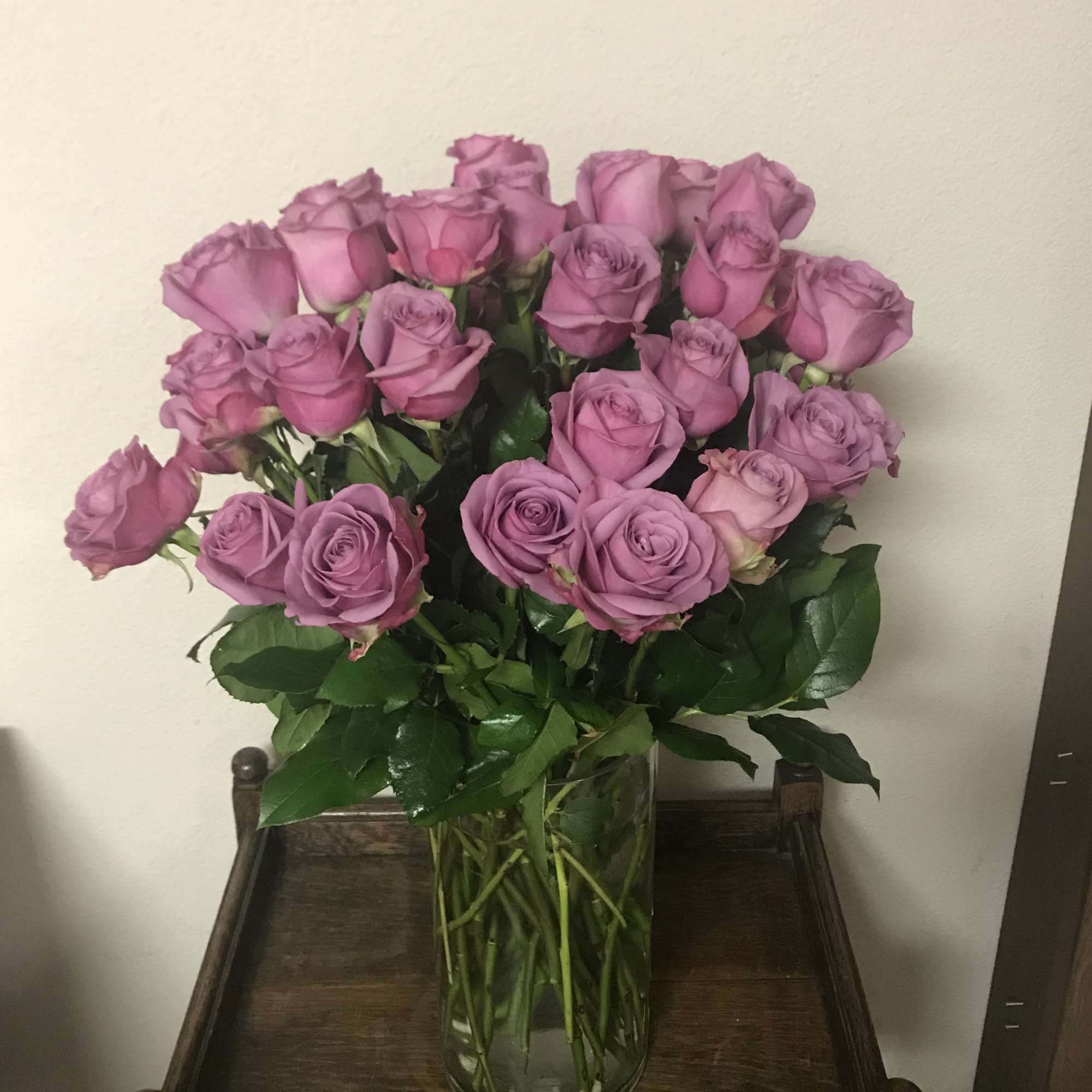 Tall clear glass vase filled with many lavender roses on a wooden table.