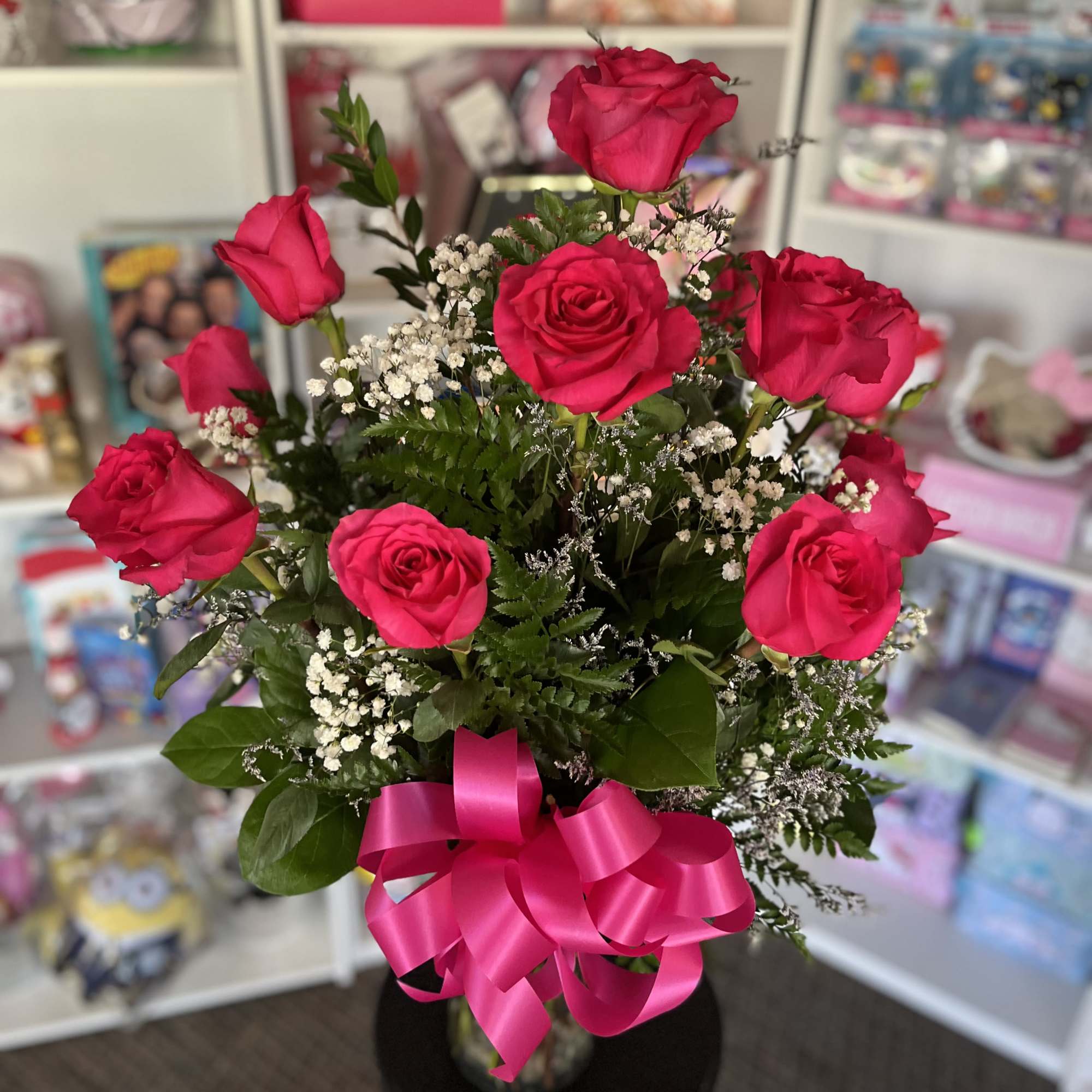 Tall arrangement of bright pink roses with white filler flowers in a glass vase with a pink bow