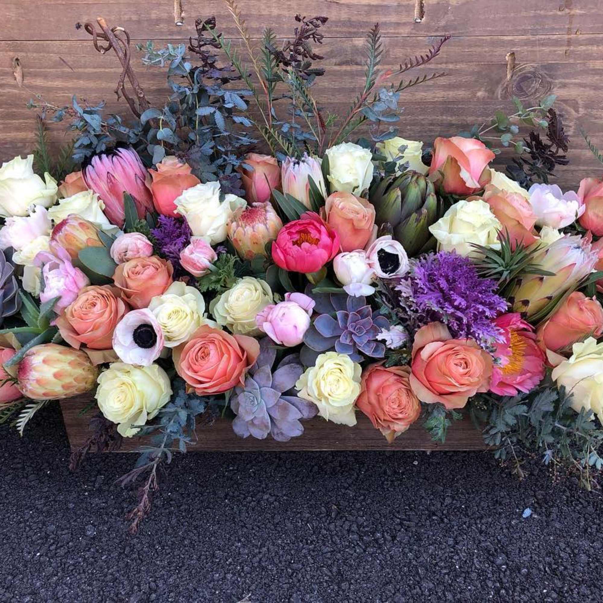 Low wooden box centerpiece with peach and cream roses, protea, anemones, succulents, and purple accents
