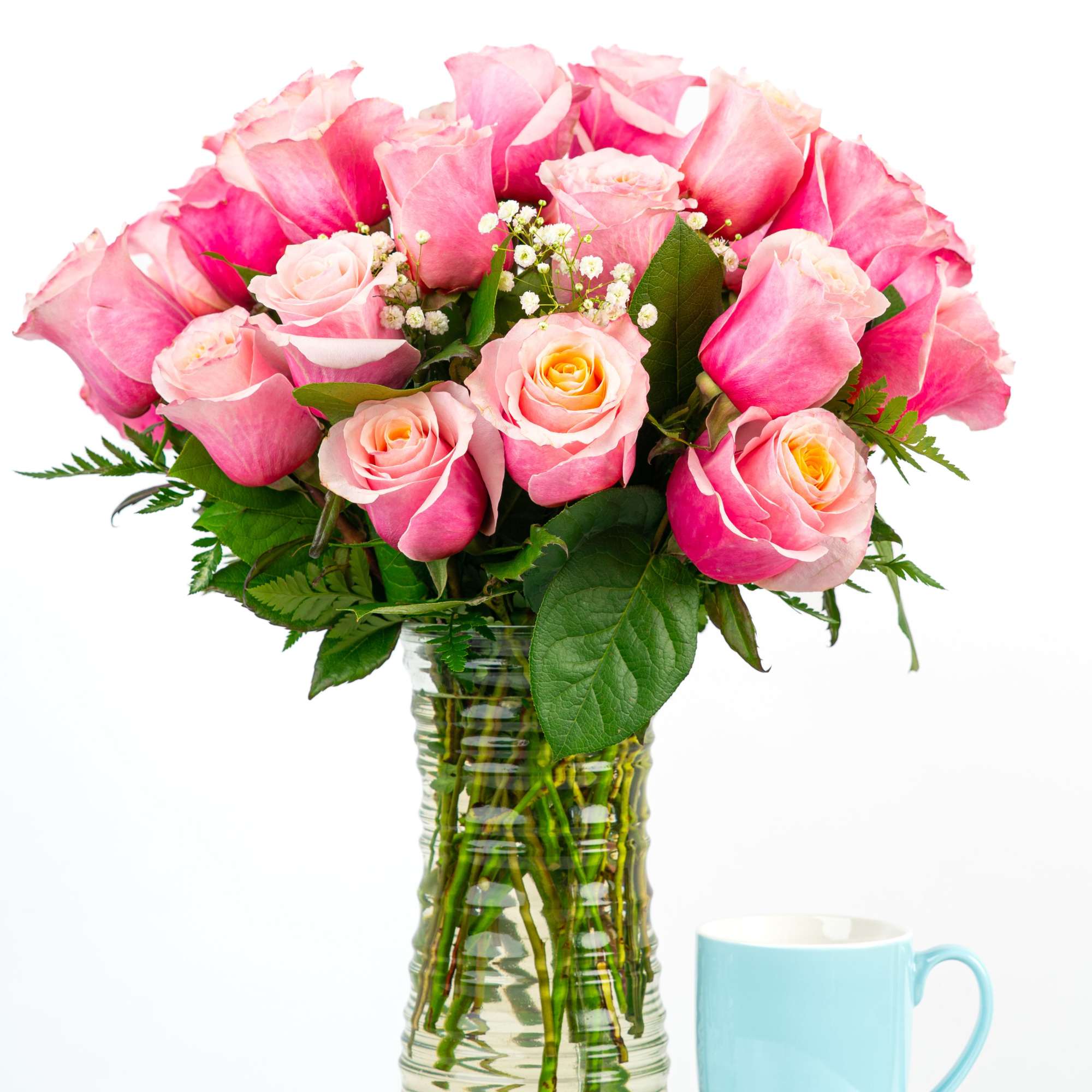 Tall bouquet of pink roses with baby's breath in a clear glass vase next to a light blue mug.