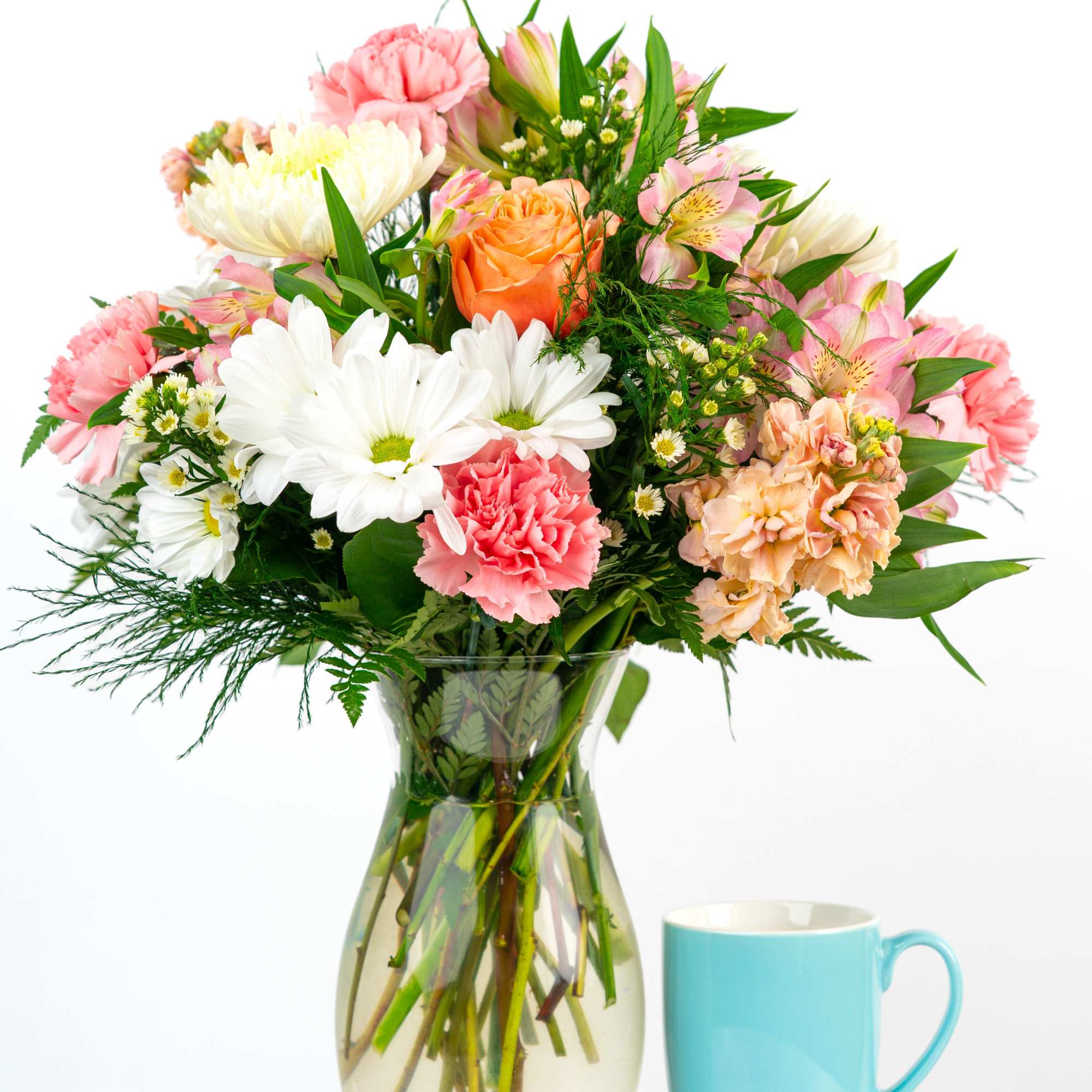 Mixed bouquet of white daisies, pink carnations, and an orange rose in a glass vase beside a blue mug