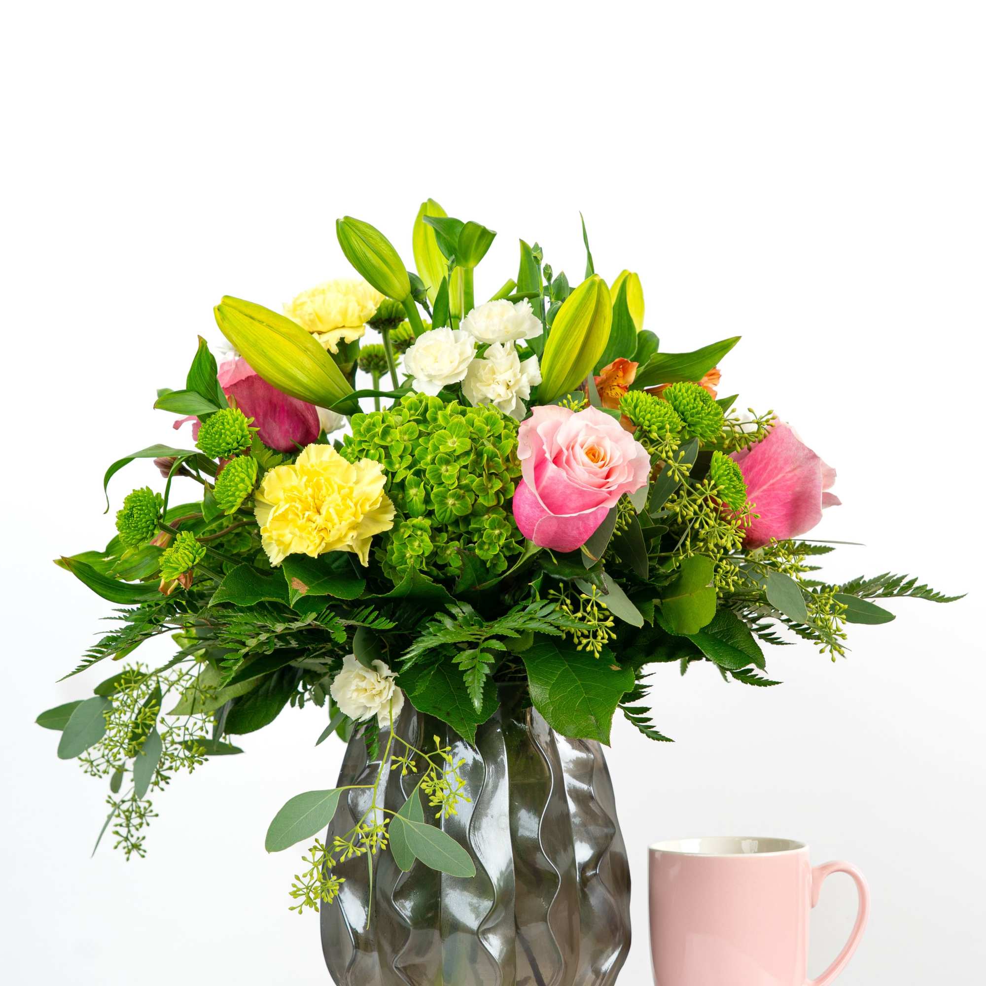 Mixed bouquet of pink roses, yellow carnations, lilies and green hydrangea in a wavy glass vase beside a pink mug