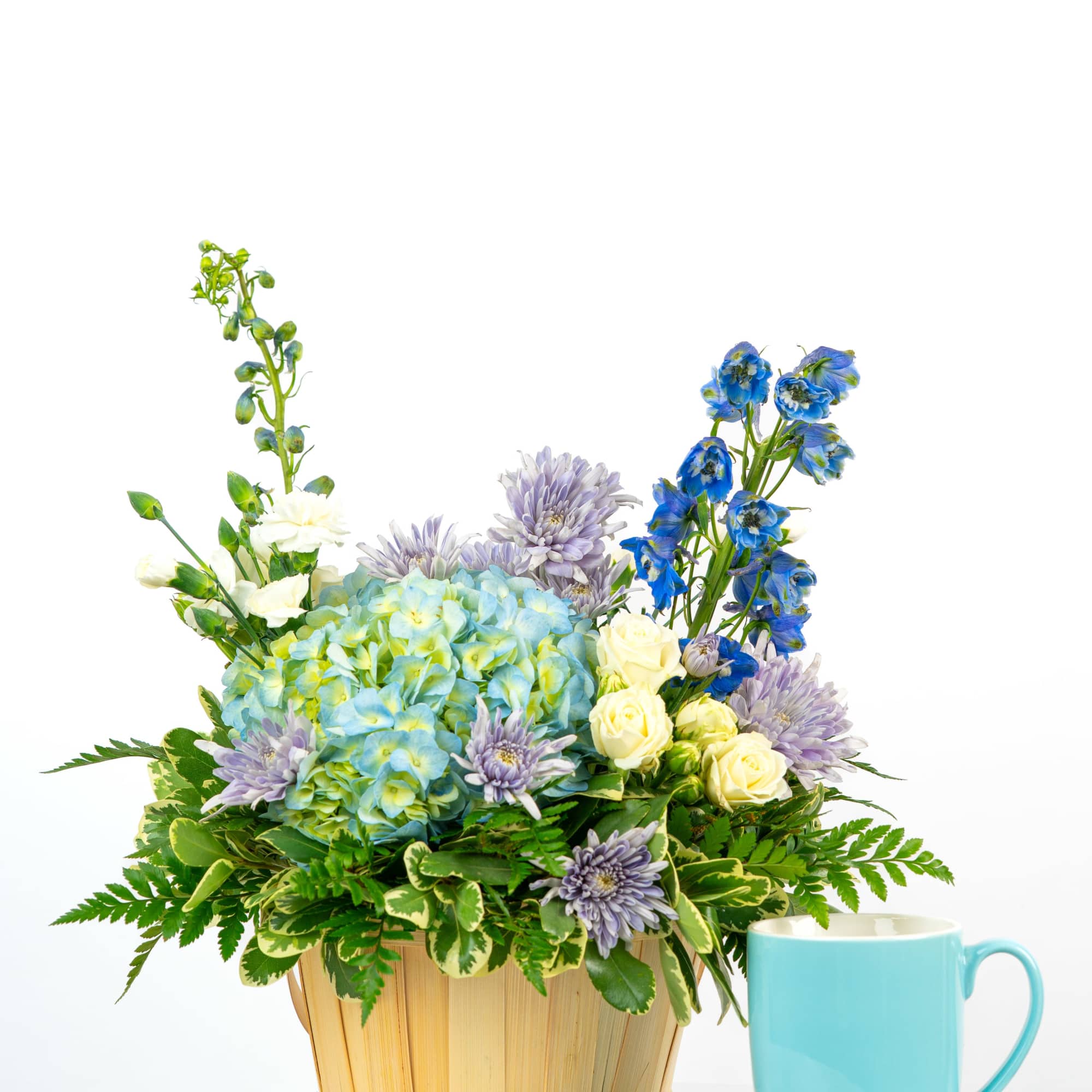 Basket arrangement of blue hydrangea, white roses, and blue blooms beside a light blue mug