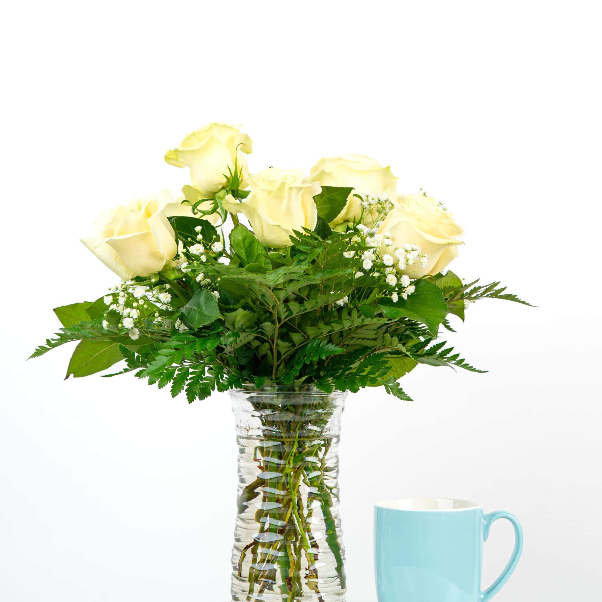 Pale yellow roses with white filler flowers in a clear glass vase beside a light blue mug