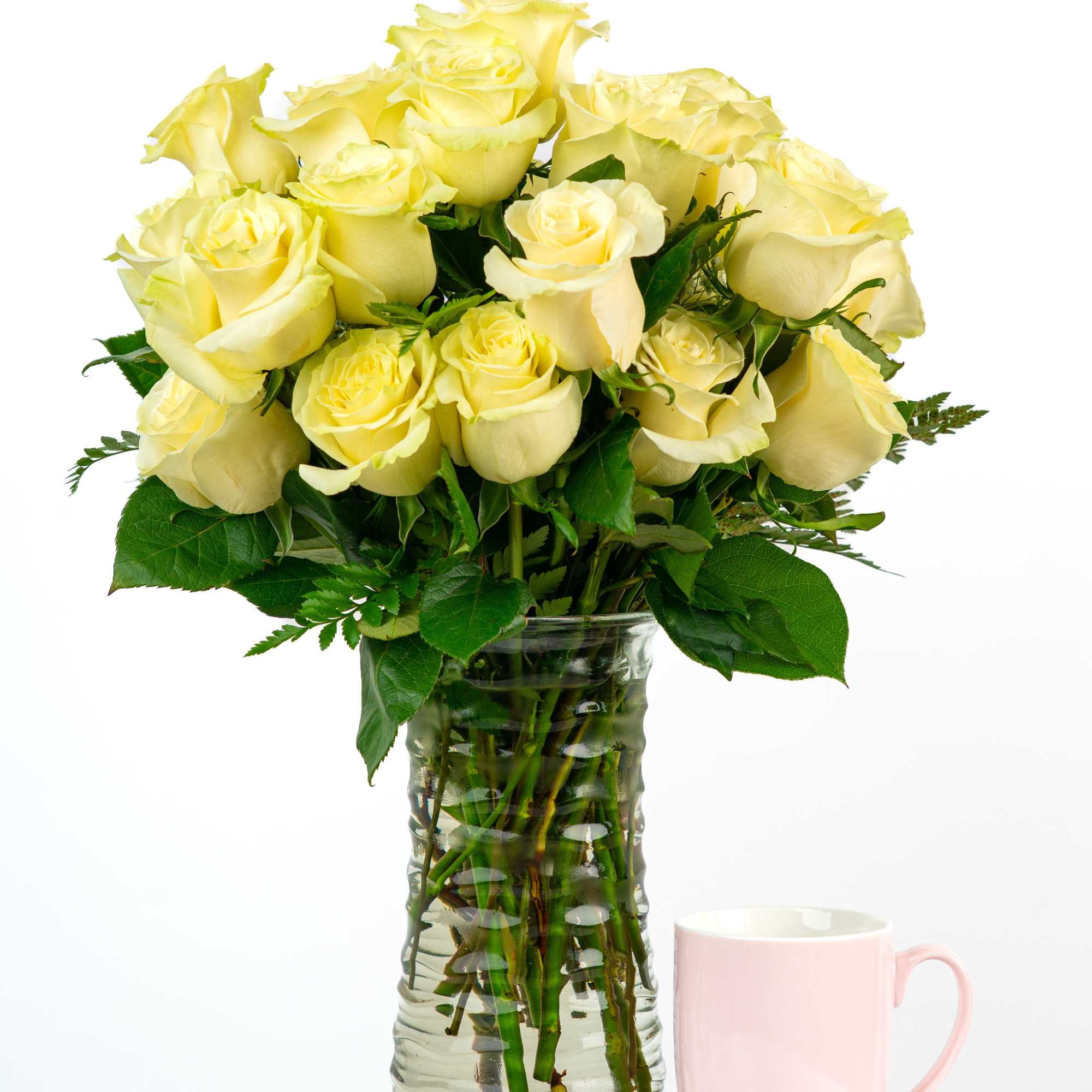 Arrangement of pale yellow roses in a tall clear glass vase beside a pink mug