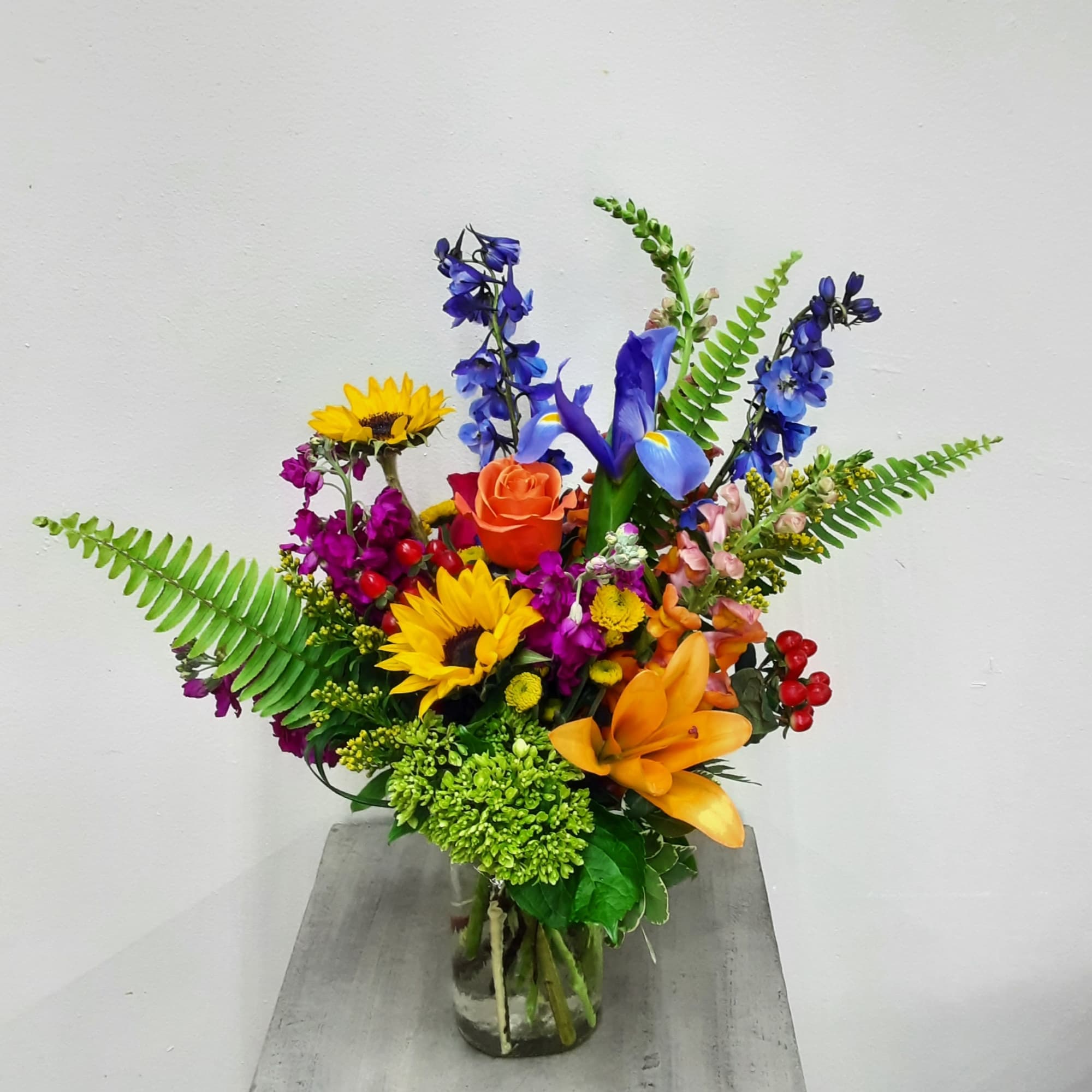 Colorful arrangement with sunflowers, orange lilies, blue delphinium, an orange rose and fern fronds in a glass vase