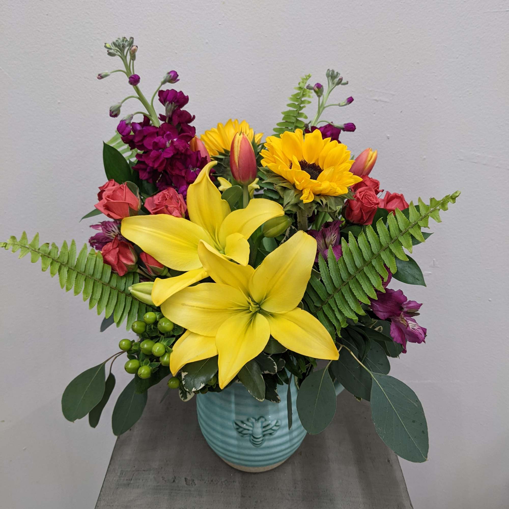 Bright arrangement of yellow lilies, sunflowers, and mixed blooms in a teal ceramic vase