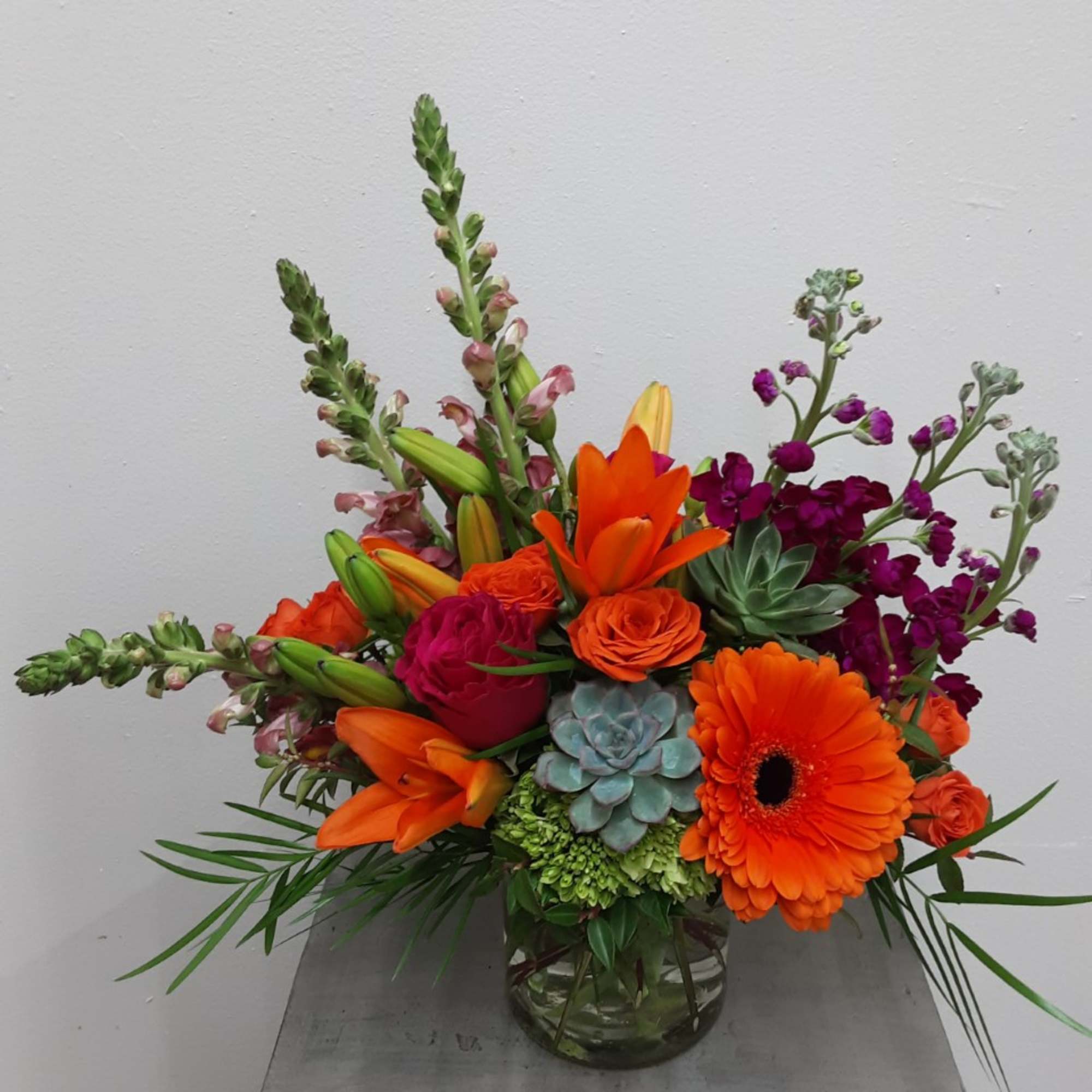 Bright orange lilies, gerbera daisy, roses, and succulents in a low clear glass vase