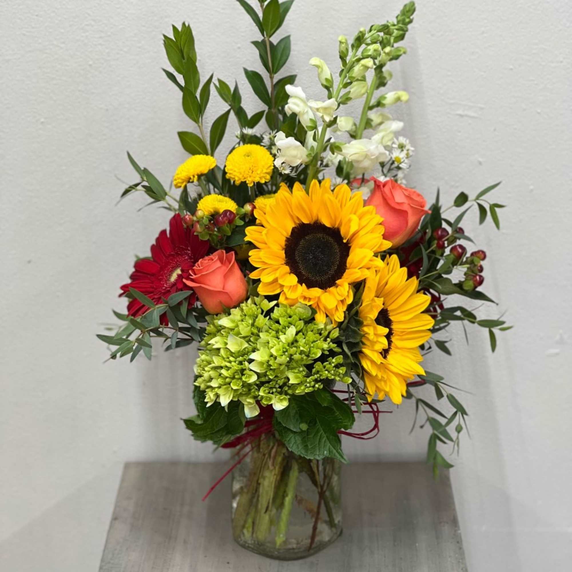 Mixed bouquet with sunflowers, orange roses, red gerberas and green hydrangea in a clear glass vase.