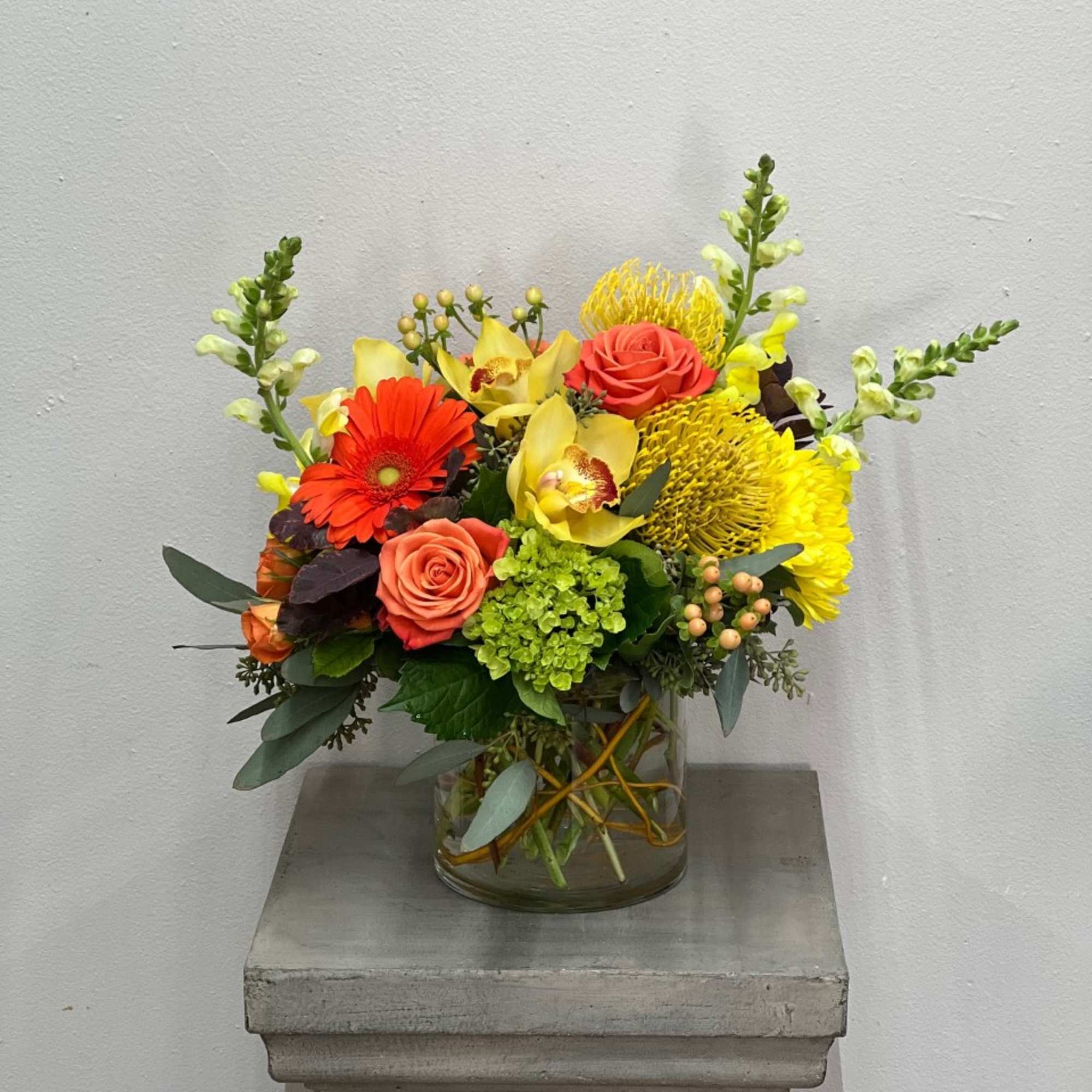 Low glass vase arrangement with orange roses, gerbera, yellow orchids, protea, and green hydrangea