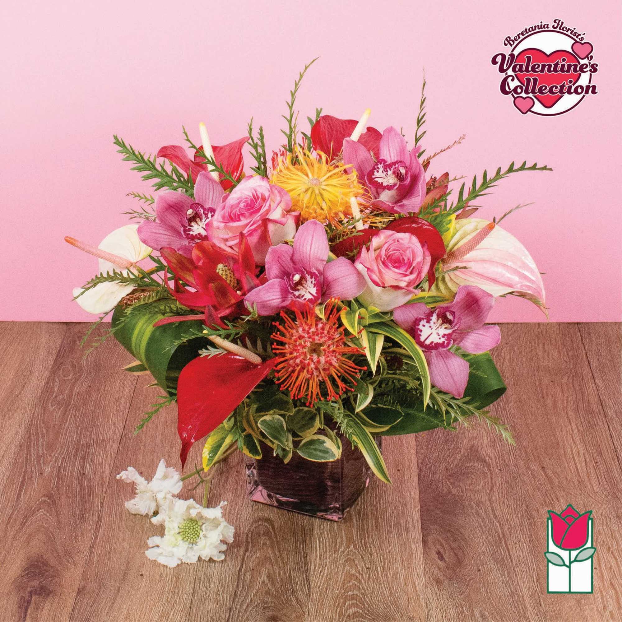 Low arrangement of pink roses, orchids, and red tropical blooms in a square glass vase on a wood surface