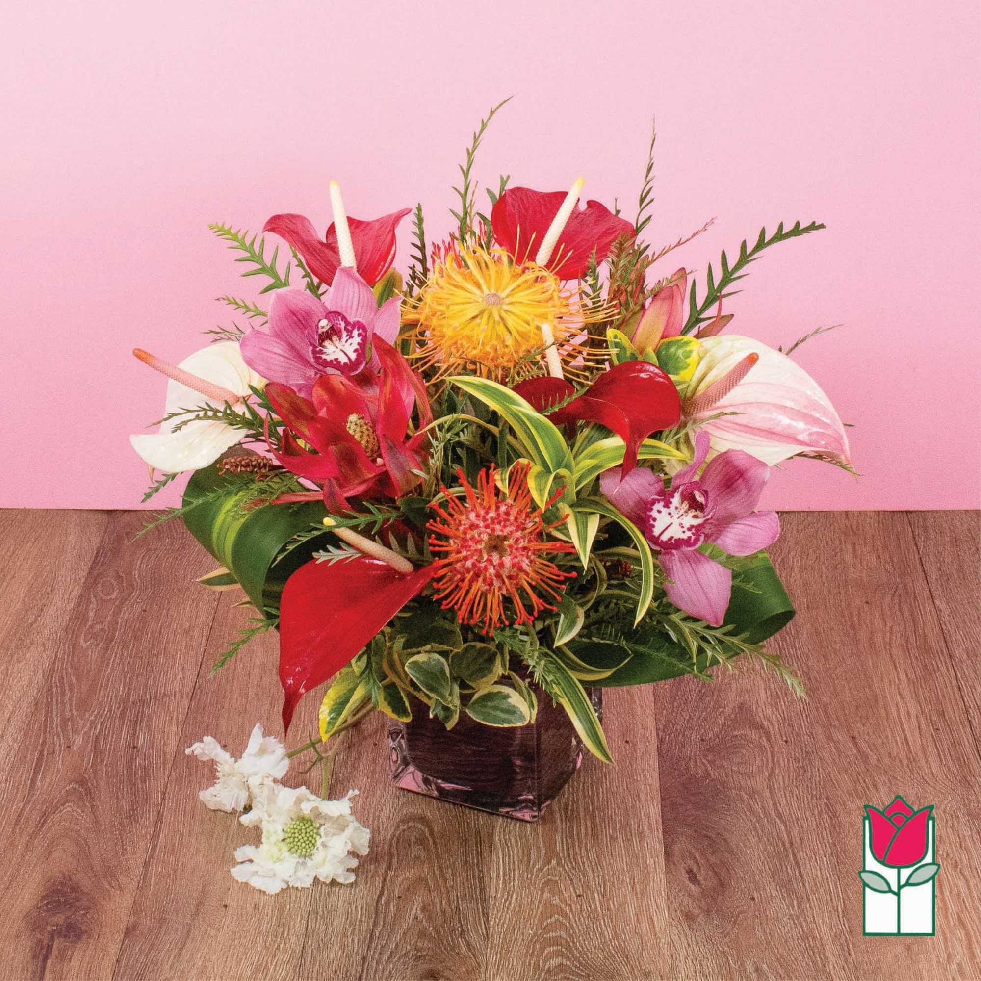 Tropical arrangement with red anthuriums, pink orchids, and yellow protea in a square glass vase.