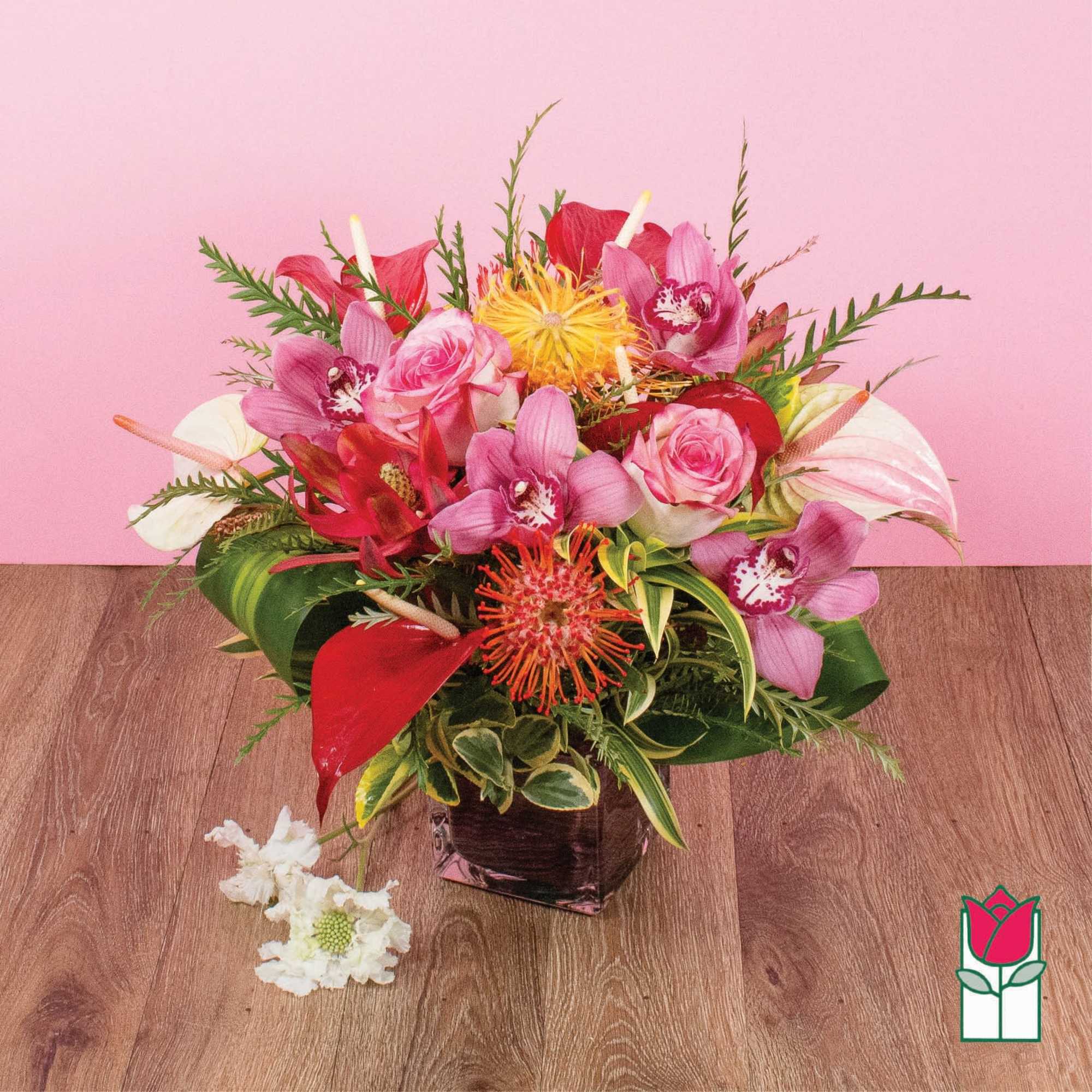 Vibrant mix of pink roses, orchids, and red tropical blooms in a square glass vase on a wood surface.